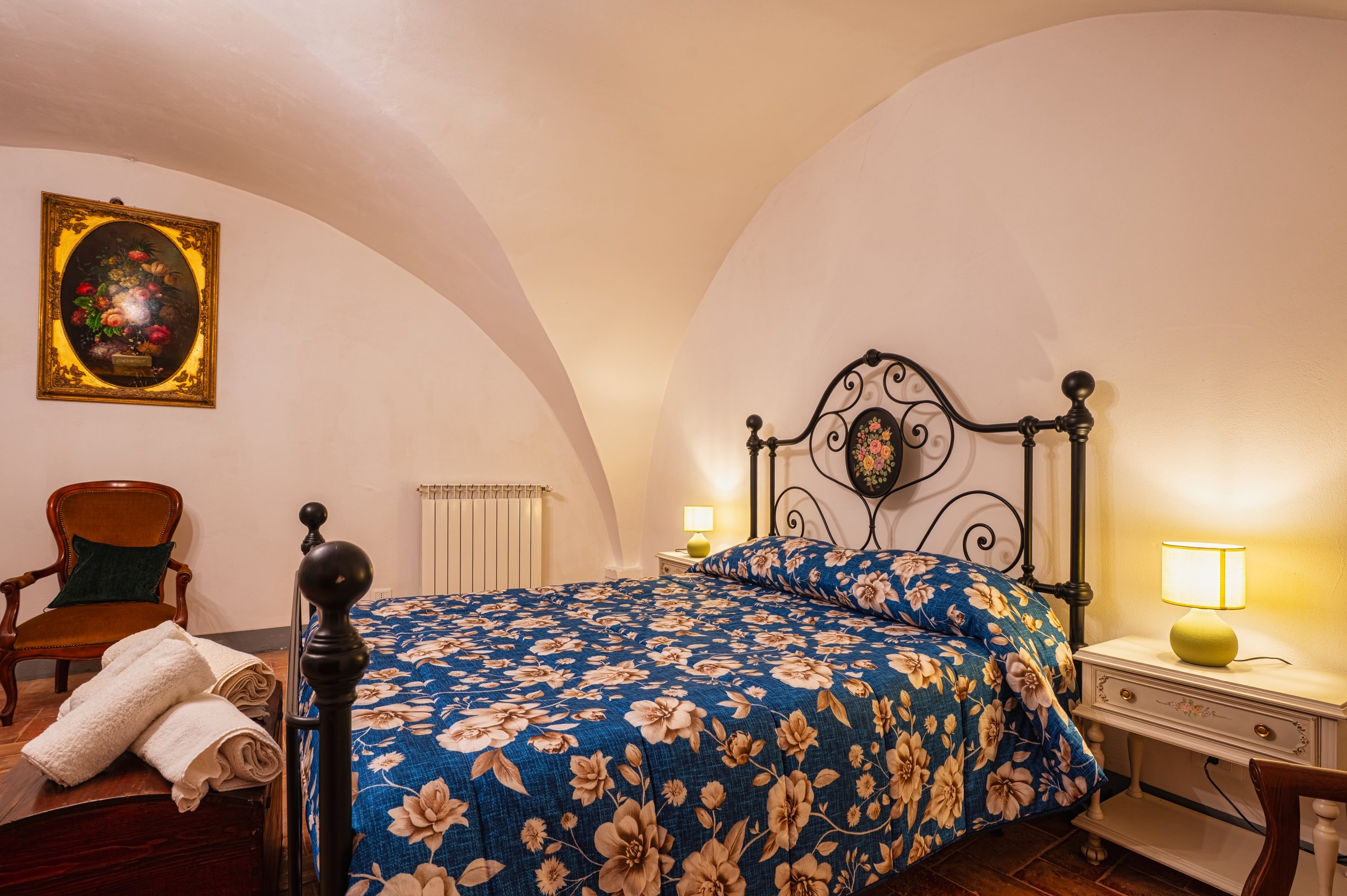 Camera da letto con letto in ferro battuto, coperte a fiori blu e beige, due lampade accese sui comodini, un quadro di fiori sulla parete e una sedia in poltrona con cuscino, alcune asciugamani su una panca in legno.