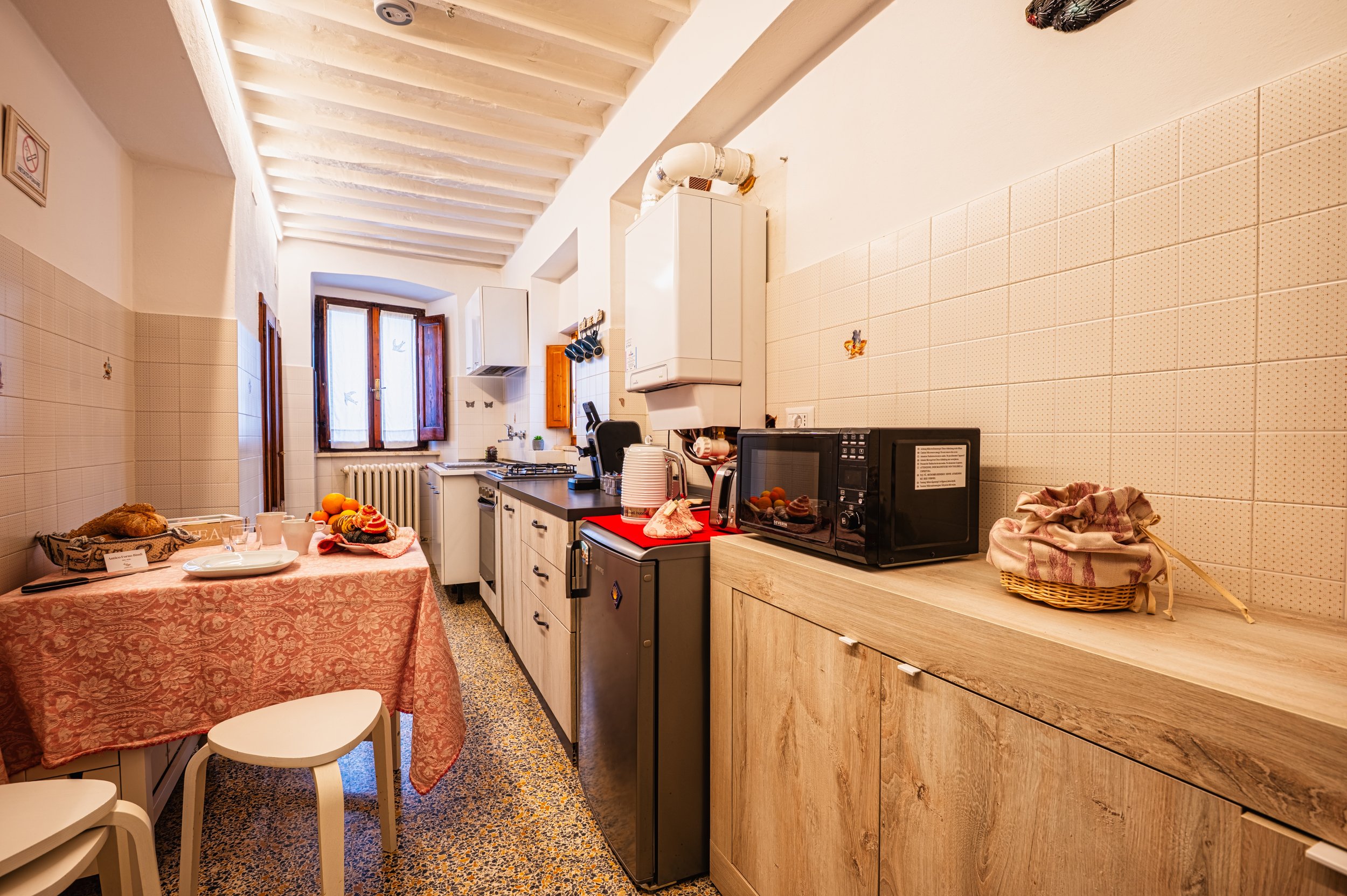 Cucina rustica con pareti in piastrelle beige, tavolo con tovaglia rosee, e vari elettrodomestici come microonde e frigorifero.