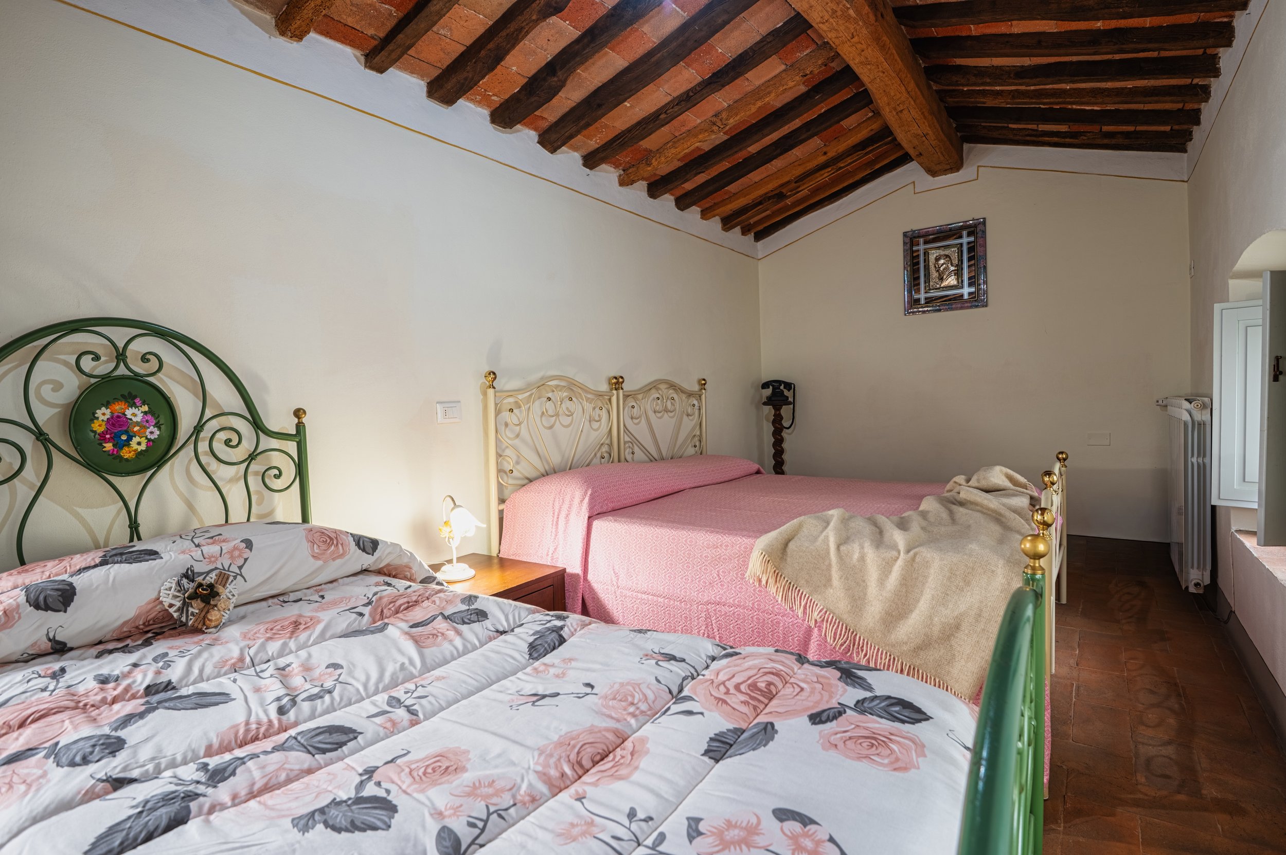 Camera da letto con due letti, uno con copripiumino a fiori rosa e uno con copriletto rosa, soffitto con travi di legno e pareti chiare. Una finestra aperta, un quadro e una lampada sul comodino.