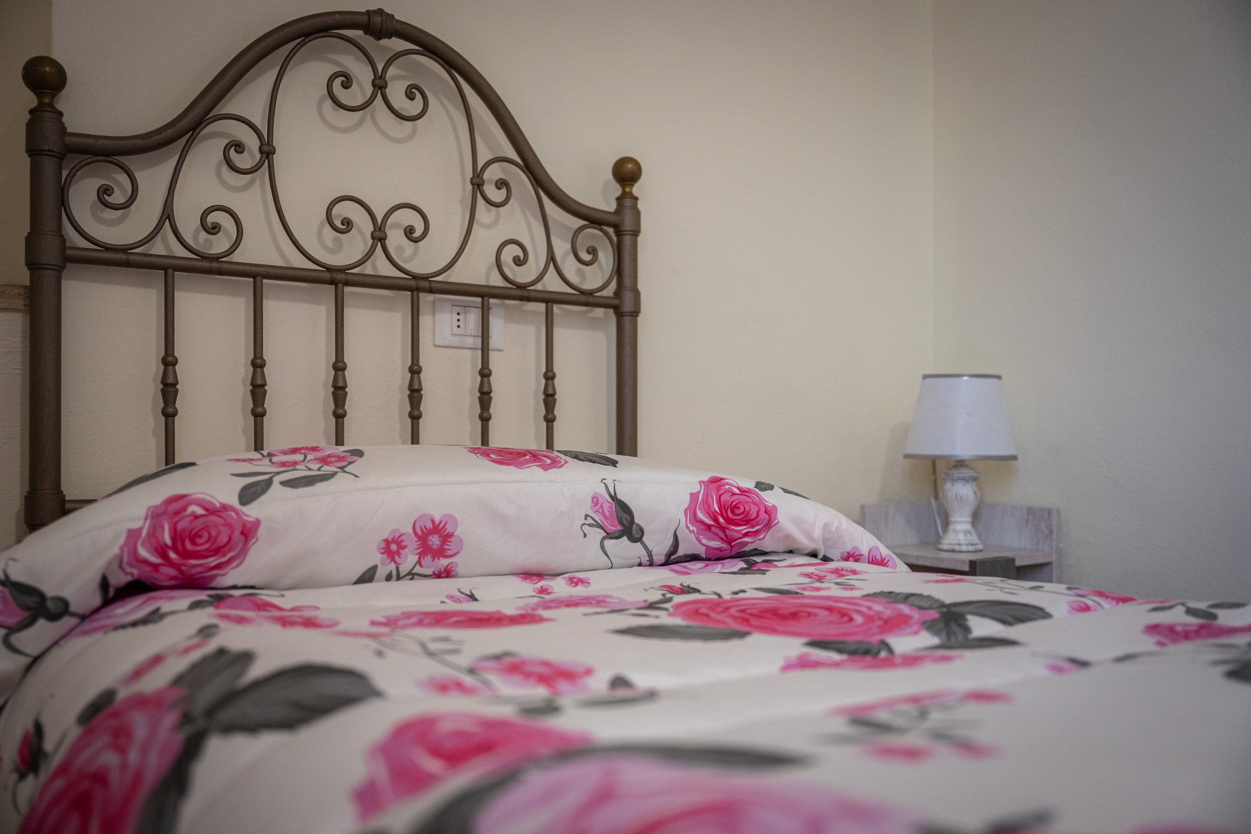 Letto matrimoniale con copripiumino e cuscino con motivo floreale rosa e grigio, lampada su comodino e parete bianca