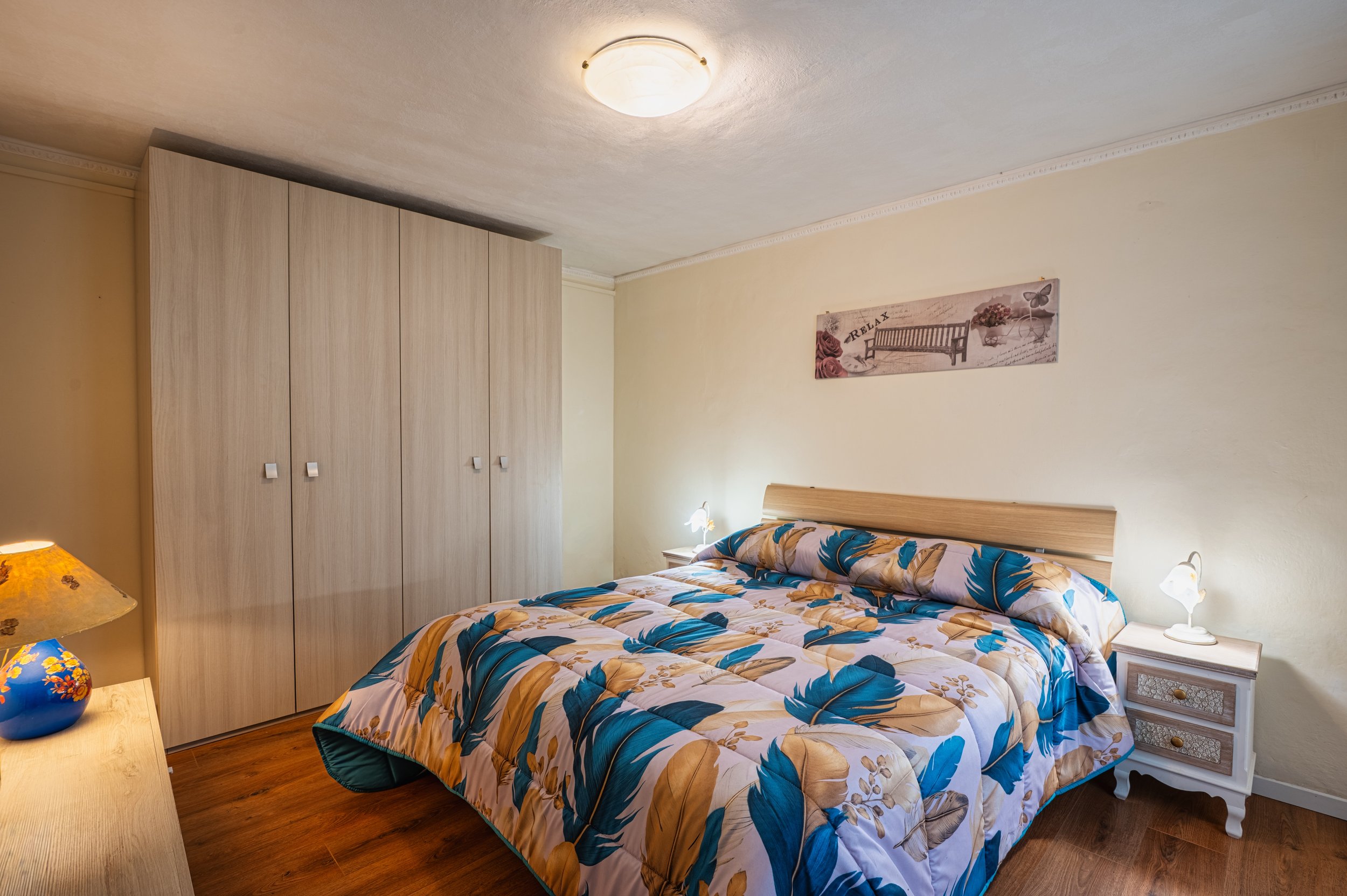 Camera da letto con letto matrimoniale, coperte con motivo di foglie verdi e beige, due lampade accanto al letto, armadio a muro in legno chiaro, pareti color crema e quadro con panchina e fiori sopra il letto.