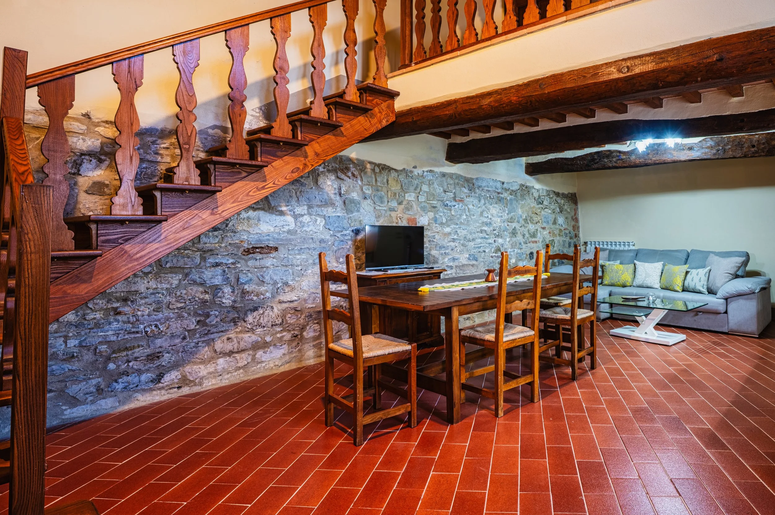 Soggiorno con parete in pietra, tavolo in legno con sei sedie, divano con cuscini di vari colori, televisore su mobile in legno, pavimento in piastrelle rosse, soffitto con travi di legno.