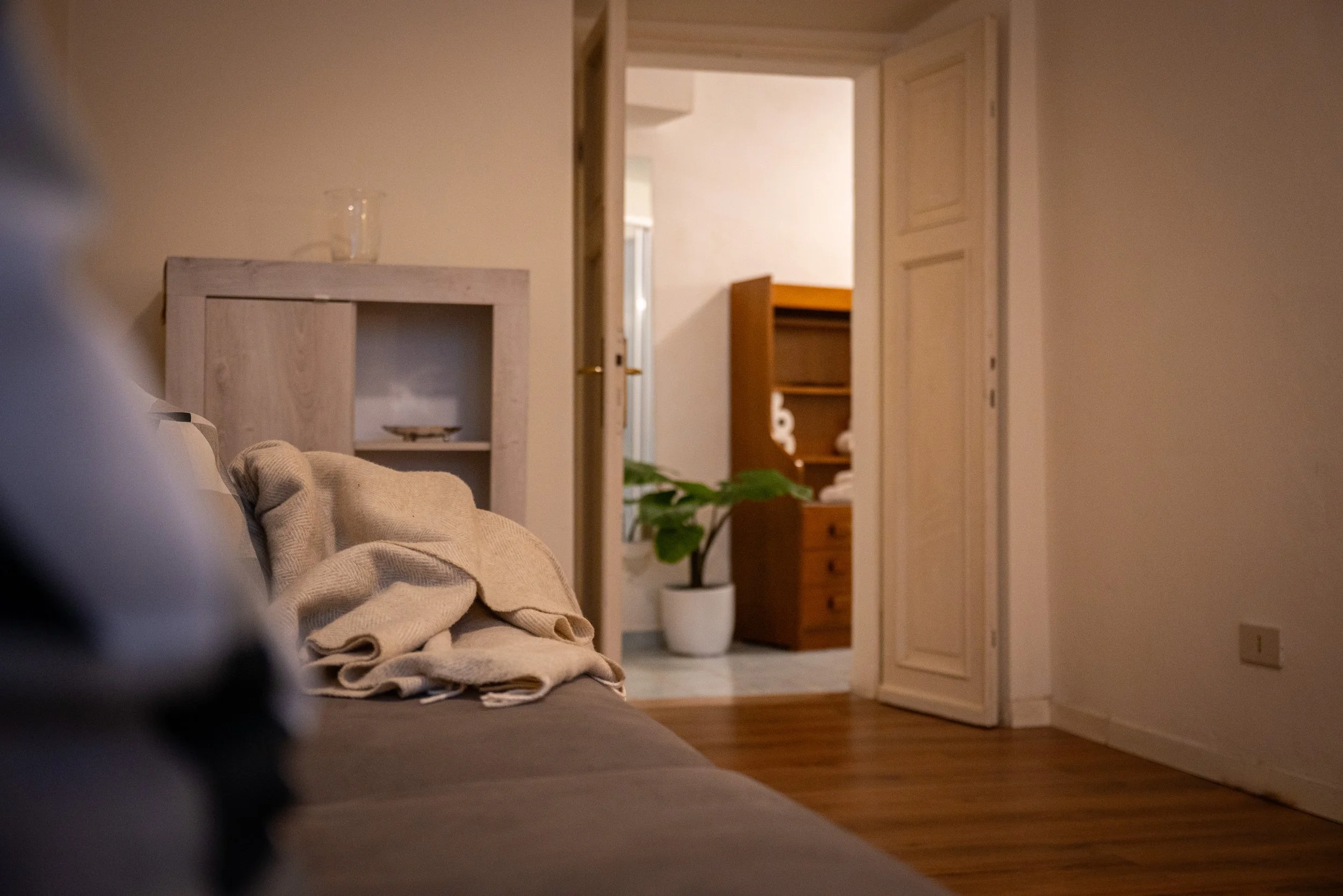 Interno di una stanza con divano, coperta beige e tavolino, vista verso una porta aperta che conduce ad un'altra stanza con una pianta in vaso e mobili in legno.