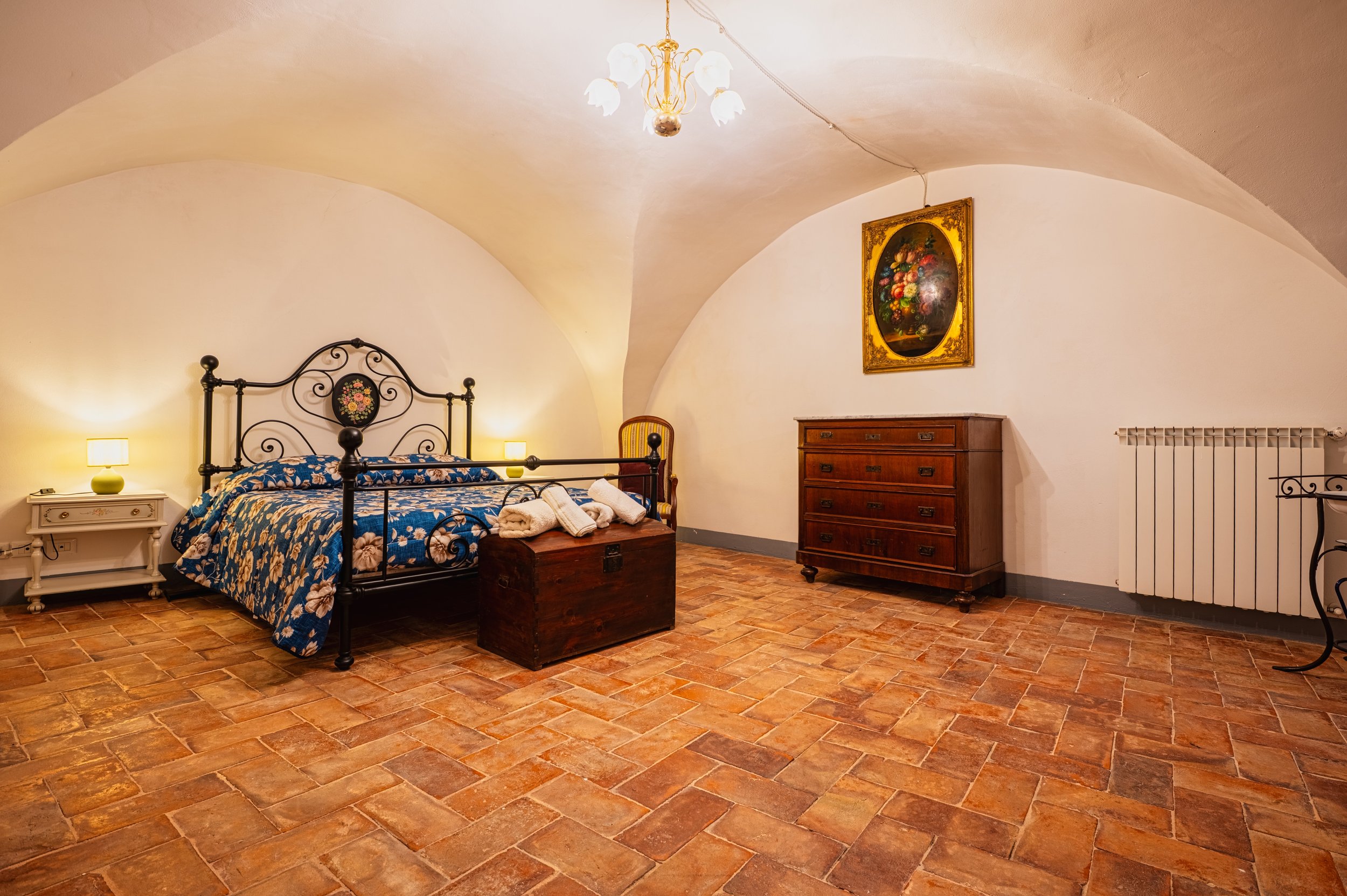 Camera da letto con soffitto a volta, letto in metallo con coperte blu con motivo floreale, due lampade da notte con lampade accese, armadio in legno, quadro con bouquet di fiori, e pavimento in piastrelle di terracotta.
