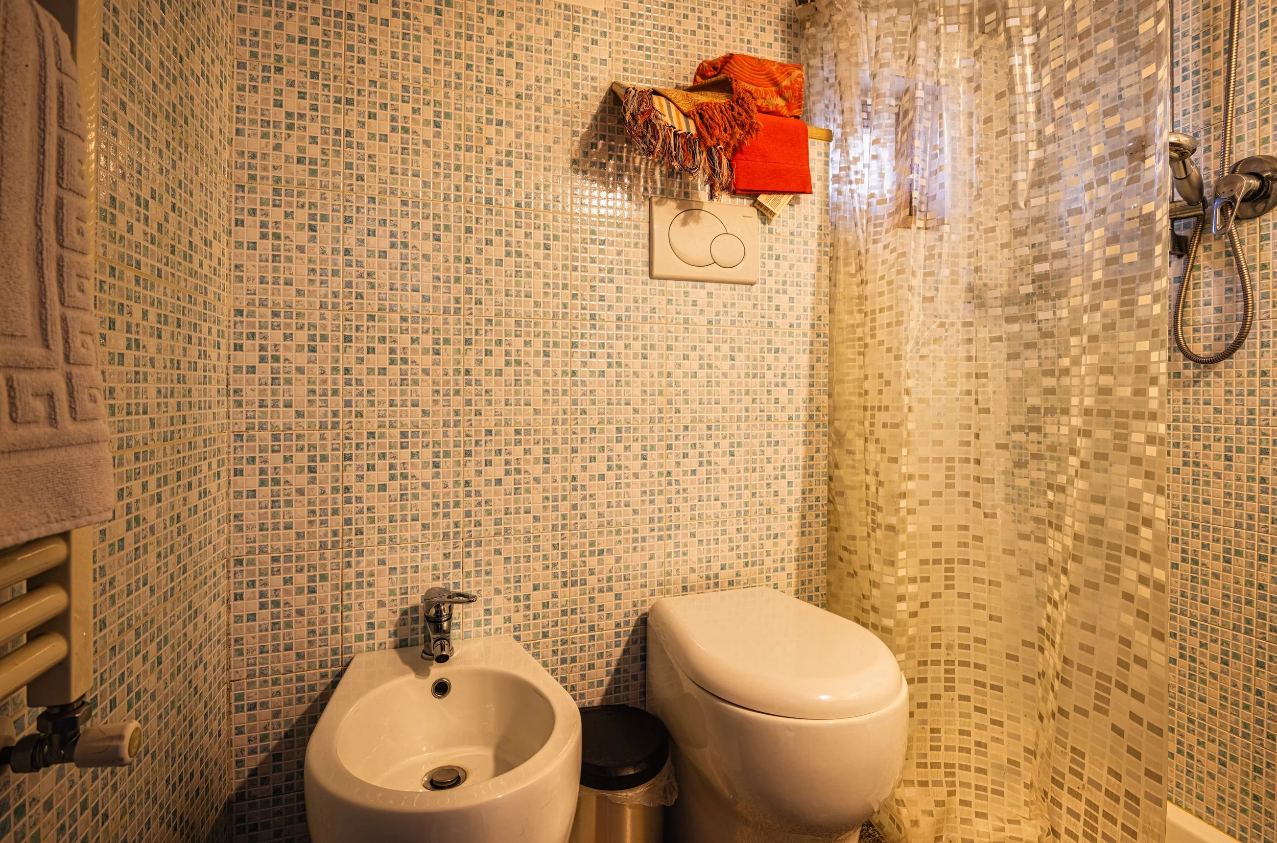 Baño con mosaico en las paredes, un inodoro de color blanco, un bidet, cortina de ducha, y una estantería con toallas y objetos.