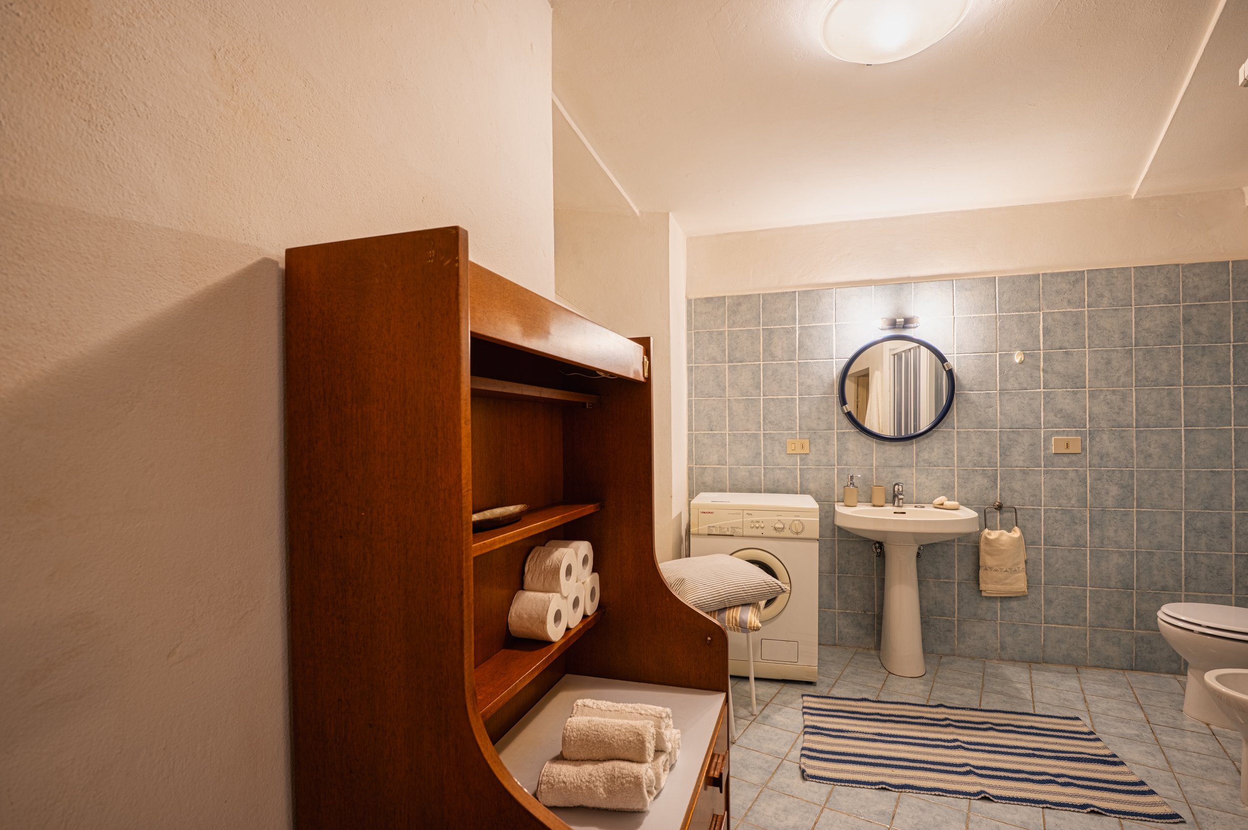 Bagno con parete rivestita di piastrelle blu, lavabo con specchio rotondo, lavatrice, asciugamano appeso e tappetino a righe, scaffale di legno con asciugamani, rotoli di carta igienica e altri oggetti.