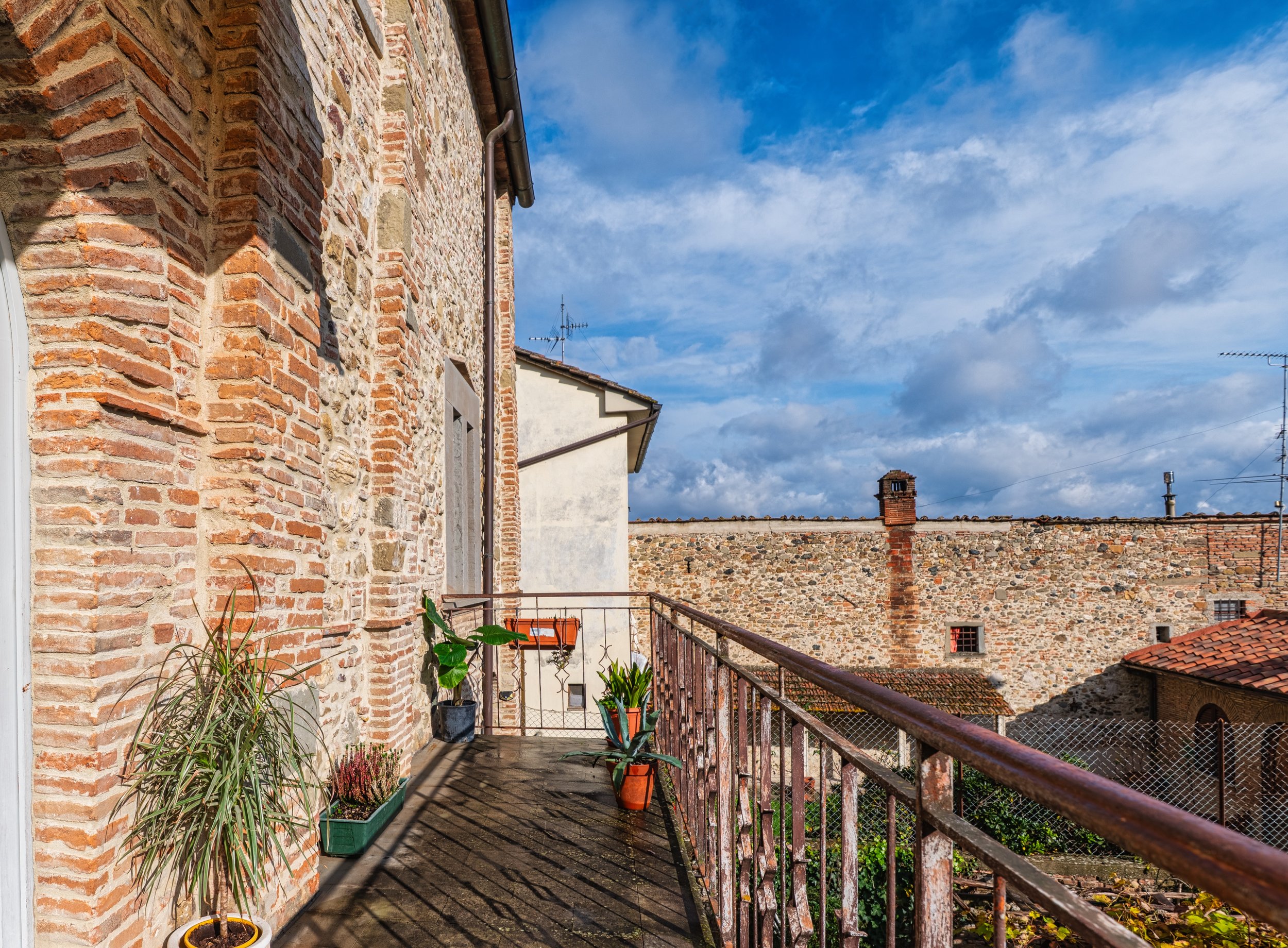 Balcone con piante in vasi variati, ringhiera di ferro, edificio in mattoni e pietra, tetti di tegole e un cielo parzialmente nuvoloso.