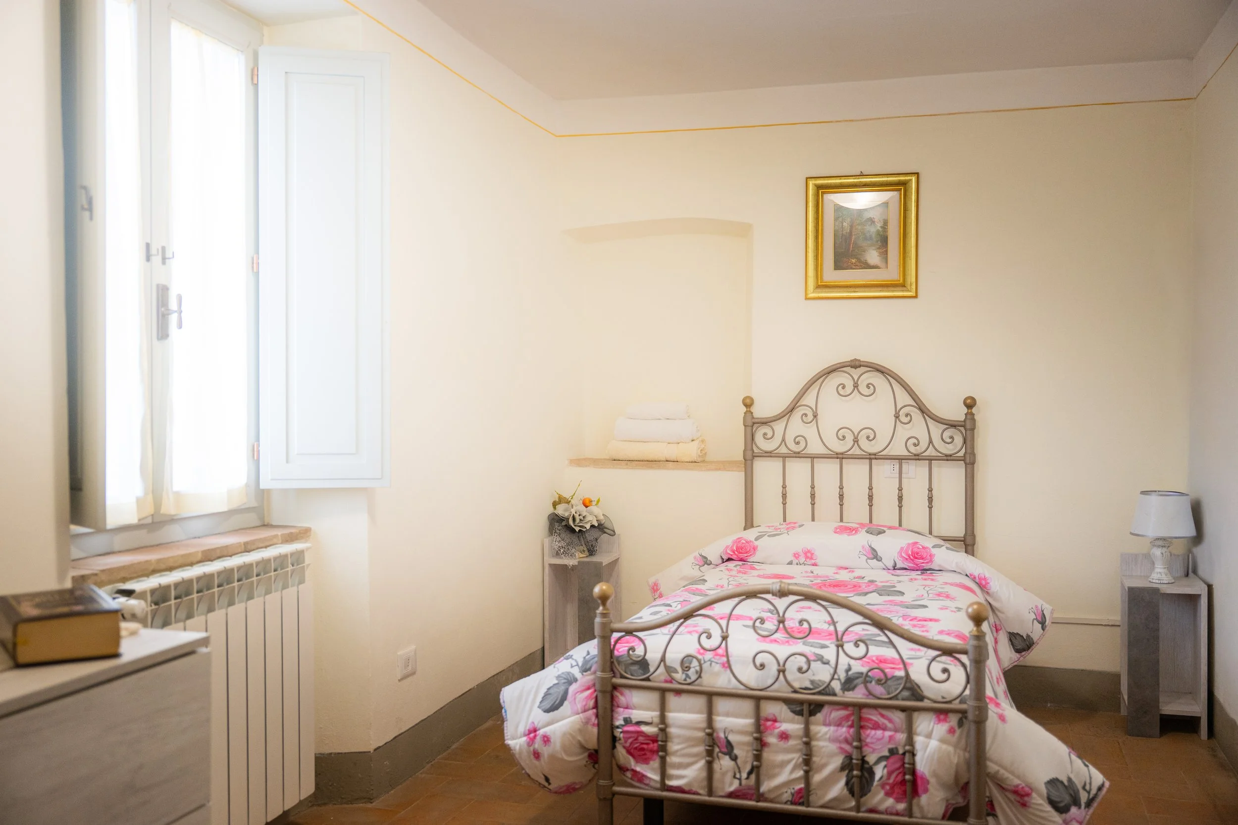 Stanza da letto con letto in ferro battuto, lenzuola con motivo floreale rosa e grigio, due comodini bianchi con lampada e piante, finestra con persiane bianche, pareti beige con cornice oro e quadro.