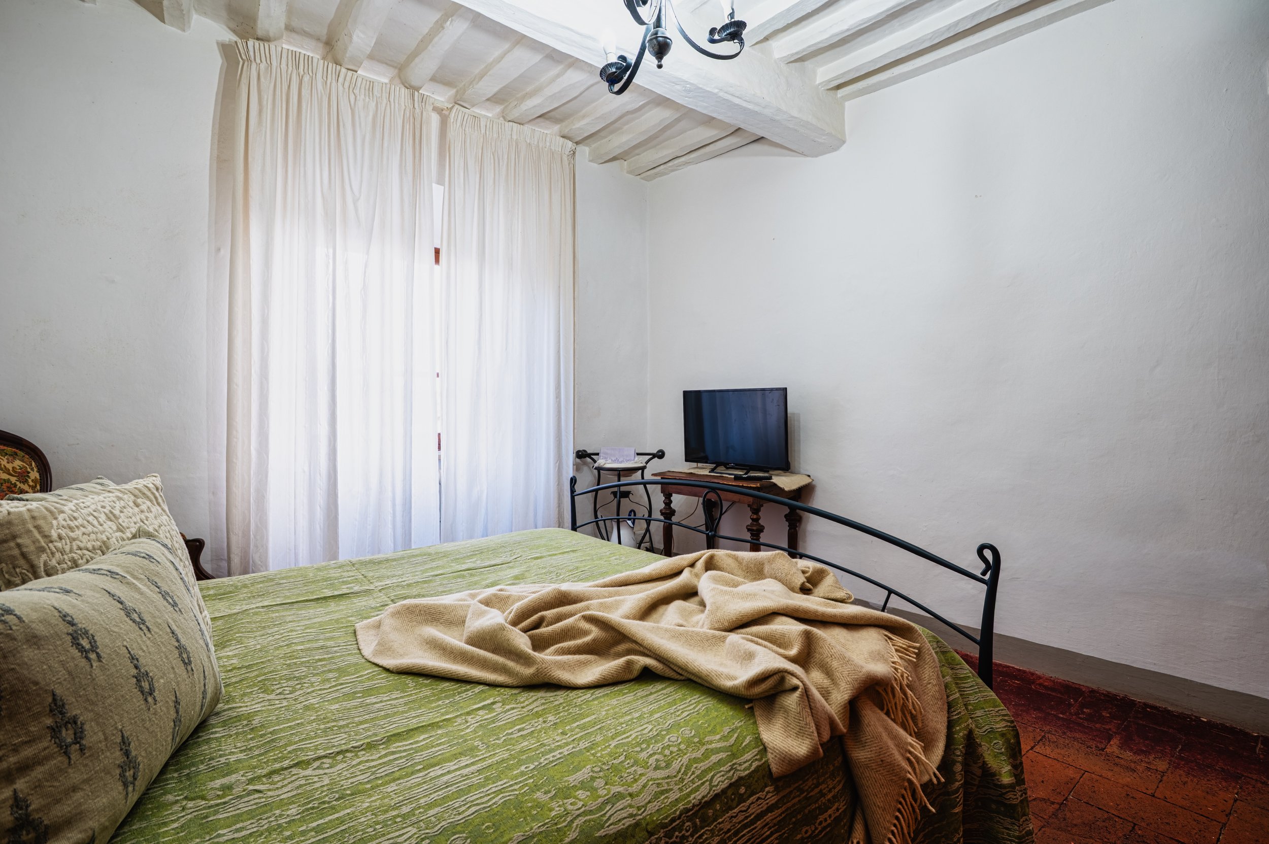 Camera in una camera da letto con letto con copripiumino verde e cuscini, lungo la parete una finestra con tende bianche, sul lato opposto un televisore su una piccola scrivania.