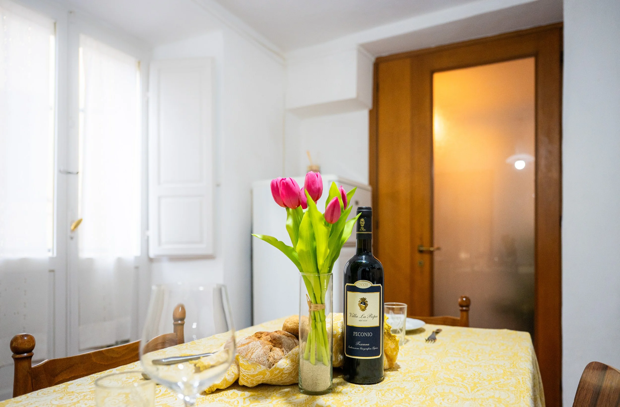 Tavola apparecchiata con un vaso di tulipani rosa, una bottiglia di vino rosso, due bicchieri e pane, in una stanza luminosa con pareti bianche, finestra con persiane e porta in legno.