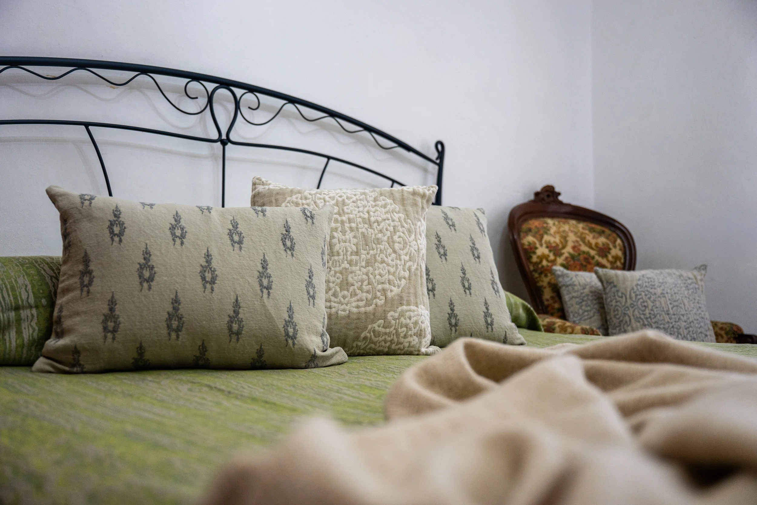 Letto con cuscini decorativi, sfondo di pareti bianche, una sedia con imbottitura floreale e altri cuscini.