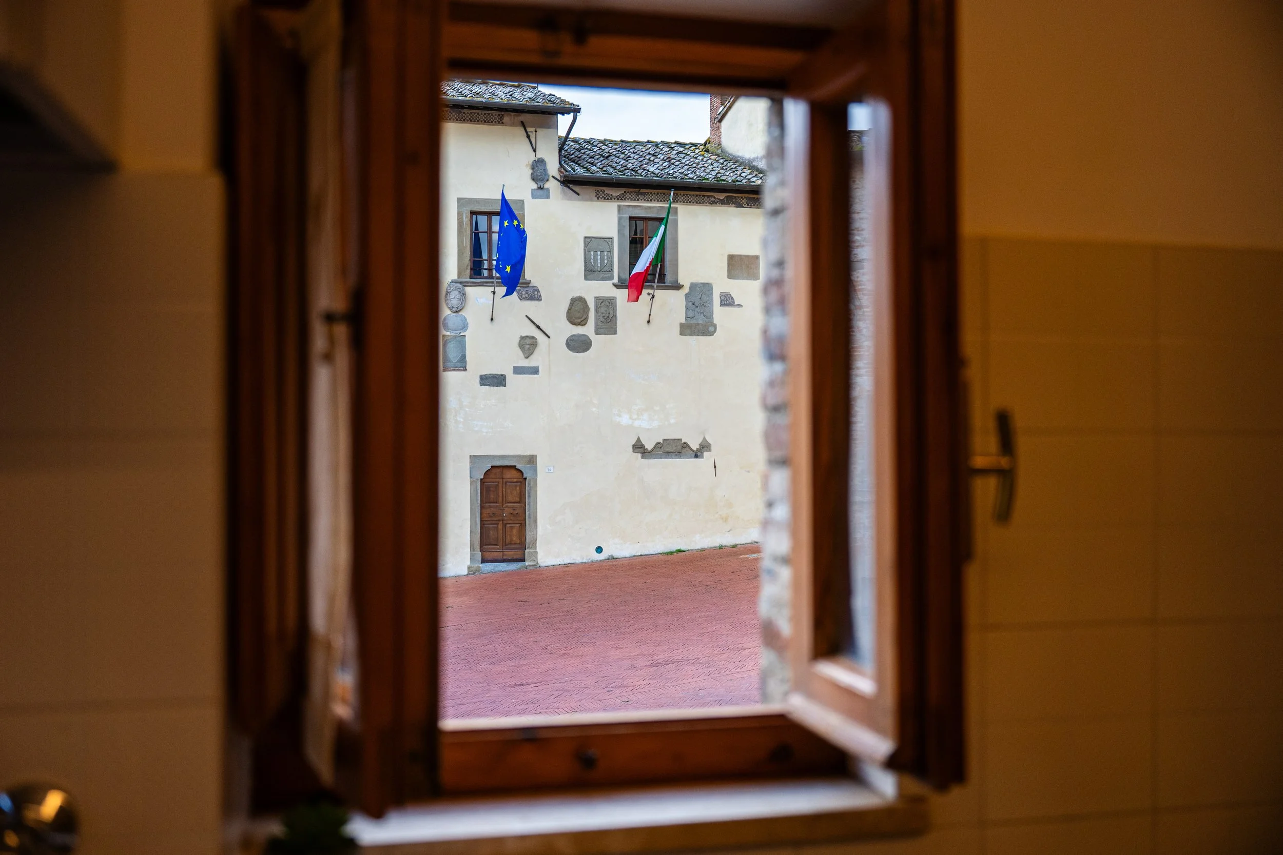 Vista di un cortile con parete decorata da stemmi e due bandiere, una dell'Unione Europea e una italiana, viste attraverso una finestra di legno.