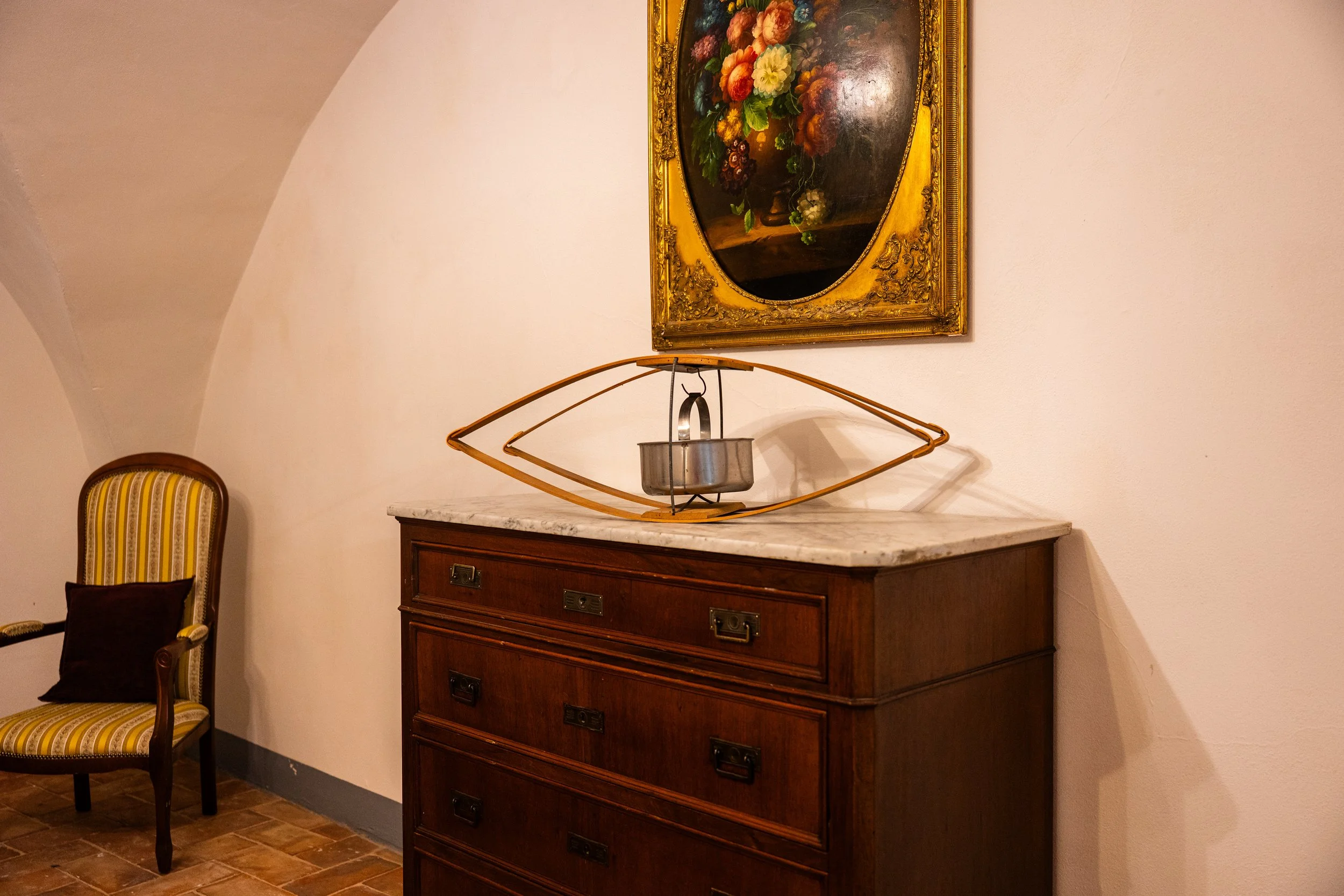 Una consolle di legno con top in marmo bianco sopra, decorata con una scultura moderna a forma di occhio stilizzato e un piatto di ciotola metallica. Sopra, un quadro con cornice dorata e dipinto di un mazzo di fiori.