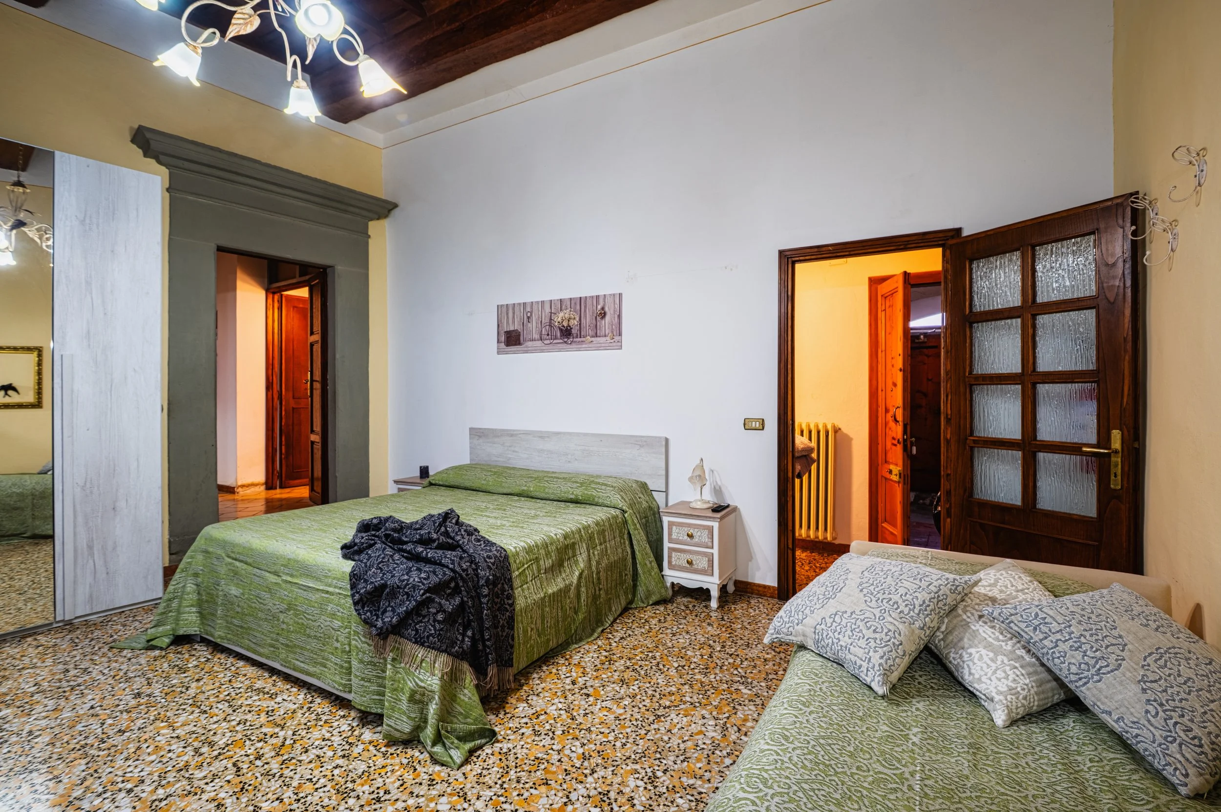 Camera da letto con due letti, uno con coperte verdi e l'altro con cuscini, pareti bianche, soffitto in legno e lampadario, armadio a specchio, porta in legno aperta che conduce a un passaggio con termosifone e altra porta di vetro