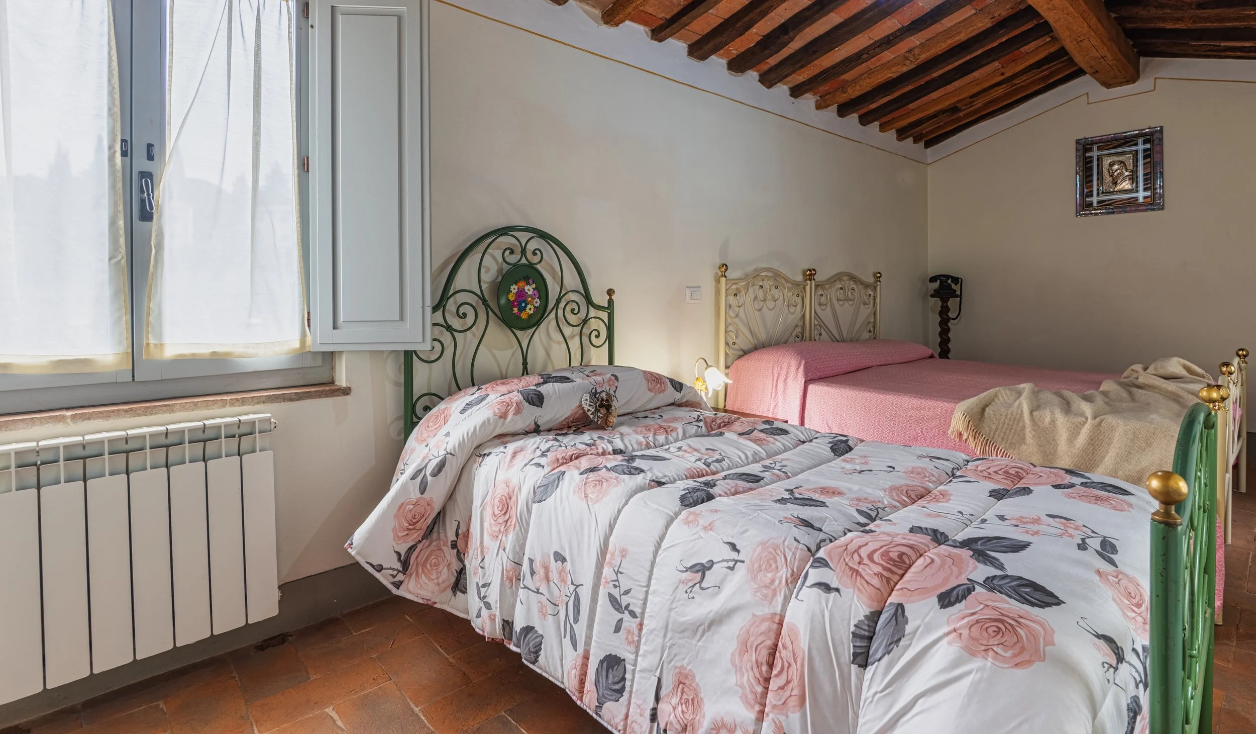 Camera da letto con due letti, uno con una coperta rosa e l'altro con una trapunta floreale, una finestra con tende bianche, soffitto in legno, pareti chiare.