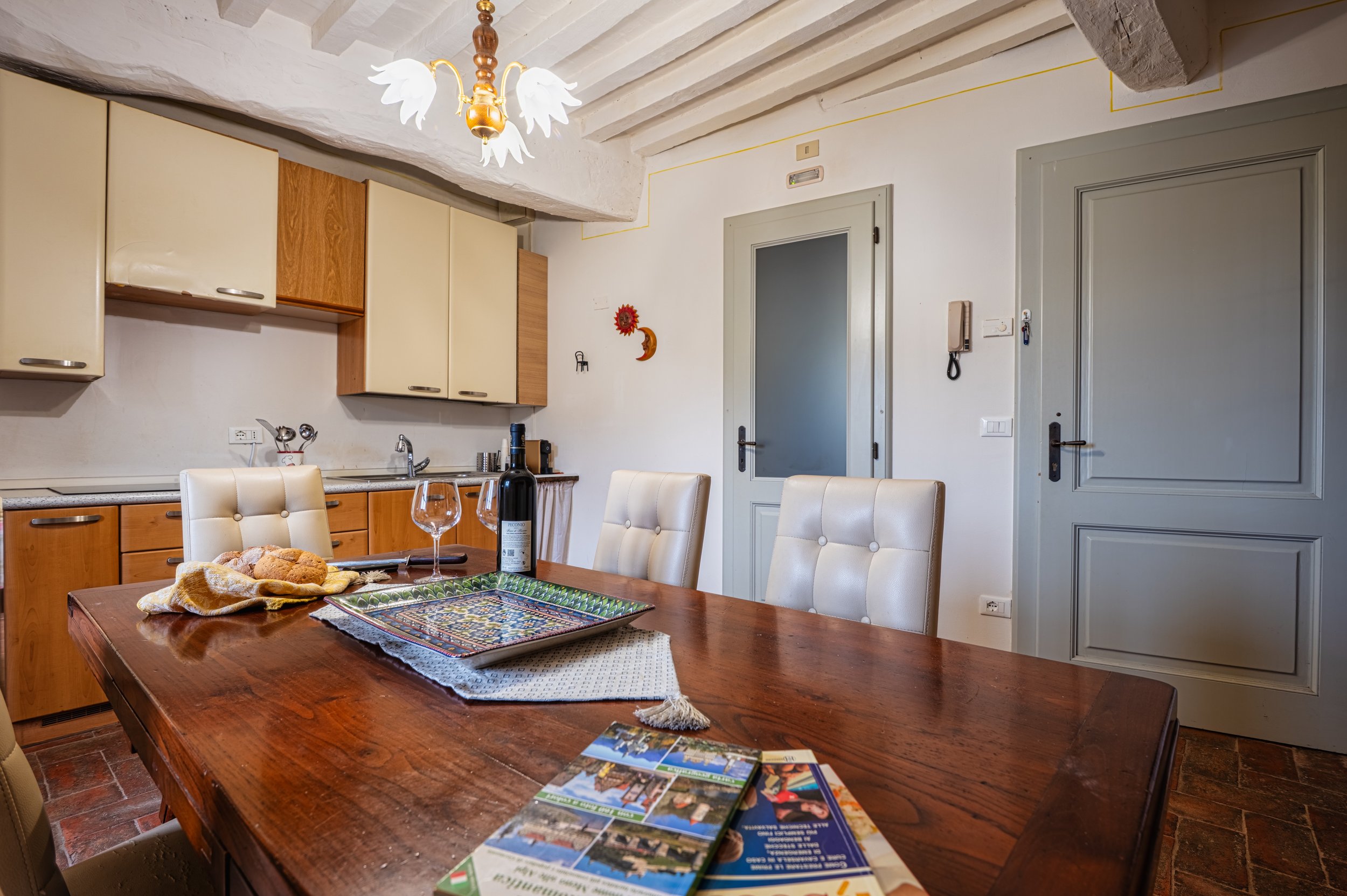 Sala da pranzo con tavolo in legno, sedie imbottite, cucina in background, luce decorativa a forma di cigno, porte grigie, bicchieri e bottiglia di vino sul tavolo.