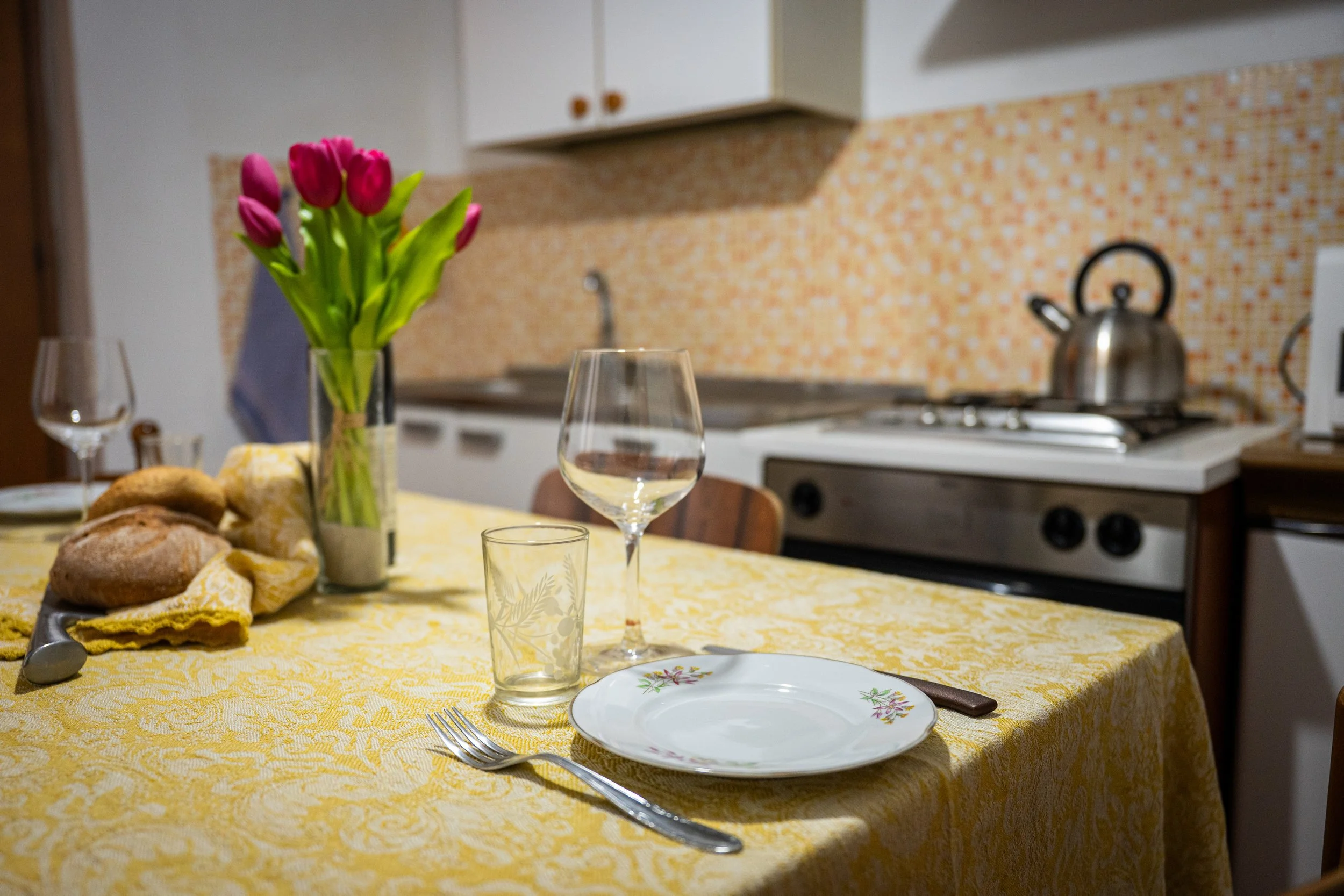 Tavolo apparecchiato con un mazzo di tulipani rosa in un vaso, due bicchieri, piatto, cucchiaio, forchetta, e pane, in una cucina con forno e pentola sul piano cottura.