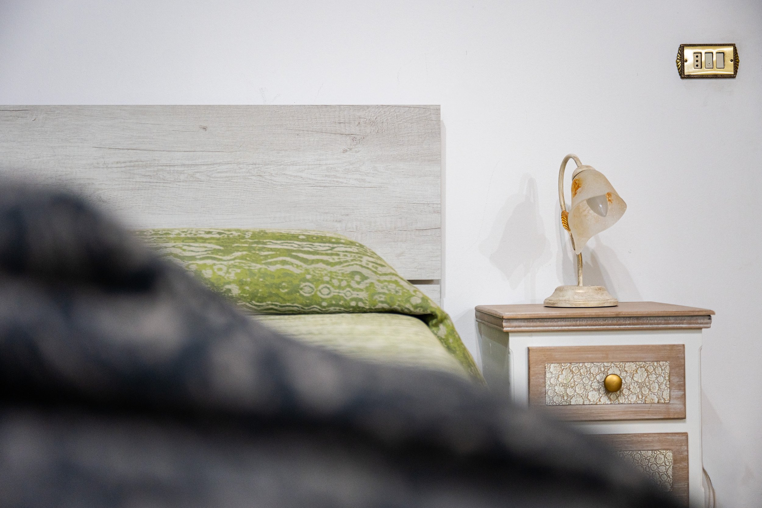 Camera da letto con letto con copriletto verde con motivo floreale, testata in legno chiaro, lampada da tavolo d'epoca sul comodino decorato, parete bianca con presa elettrica a muro.