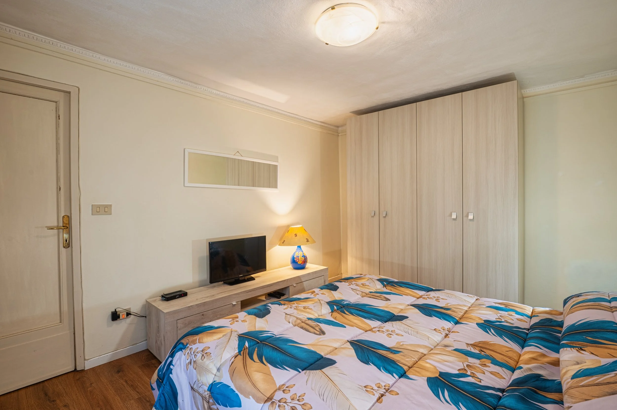 Camera da letto con letto matrimoniale, armadio a muro, televisore, lampada colorata, pareti beige, soffitto con luce rotonda, pavimento in legno