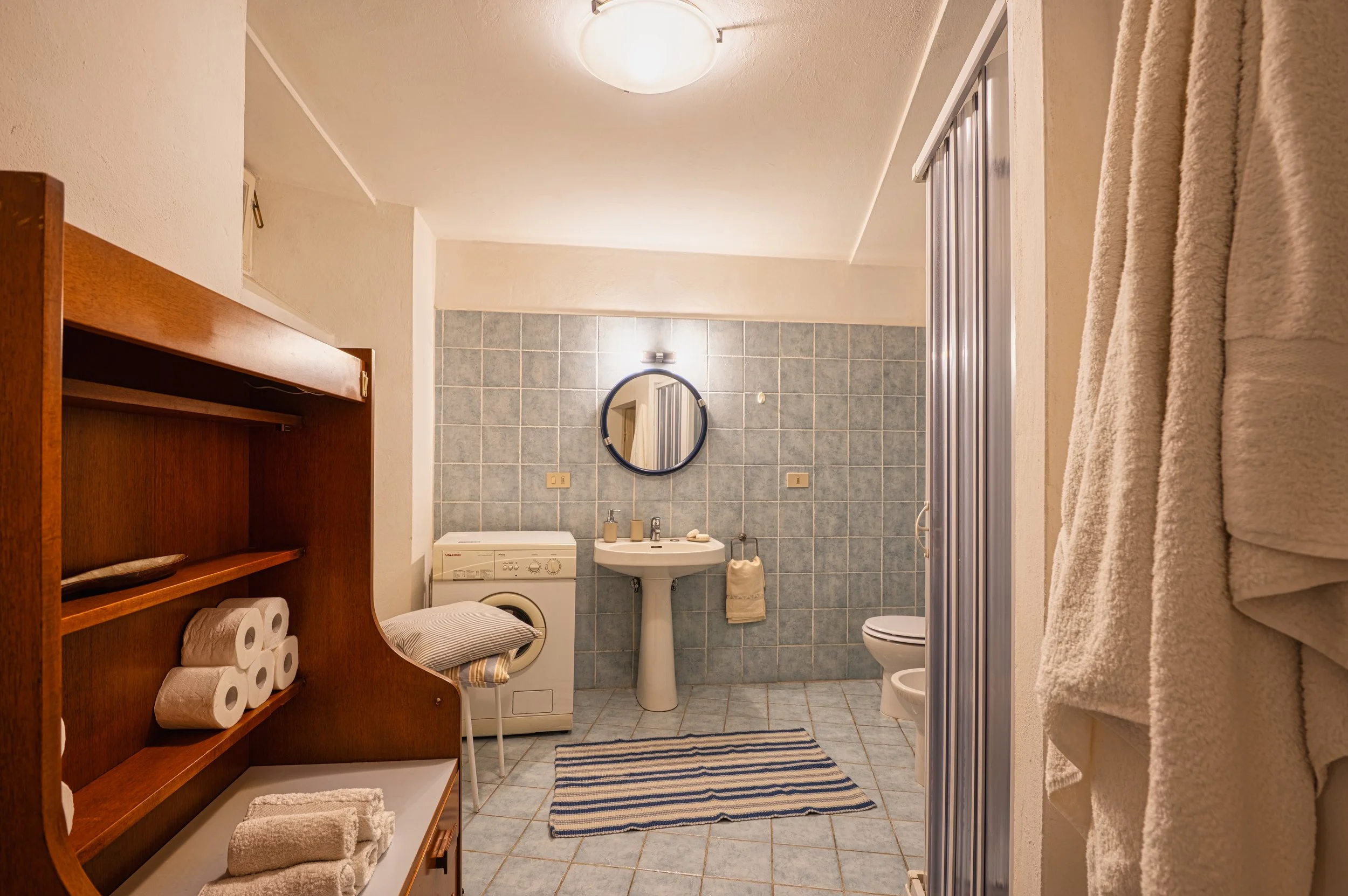 Bagno con piastrelle azzurre, presenza di lavabo, specchio rotondo, toilette, asciugamani, lavatrice, armadietto di legno con rotoli di carta, apparecchio di illuminazione sopra lo specchio, porta della doccia, tappeto a righe sulla pavimentazione.