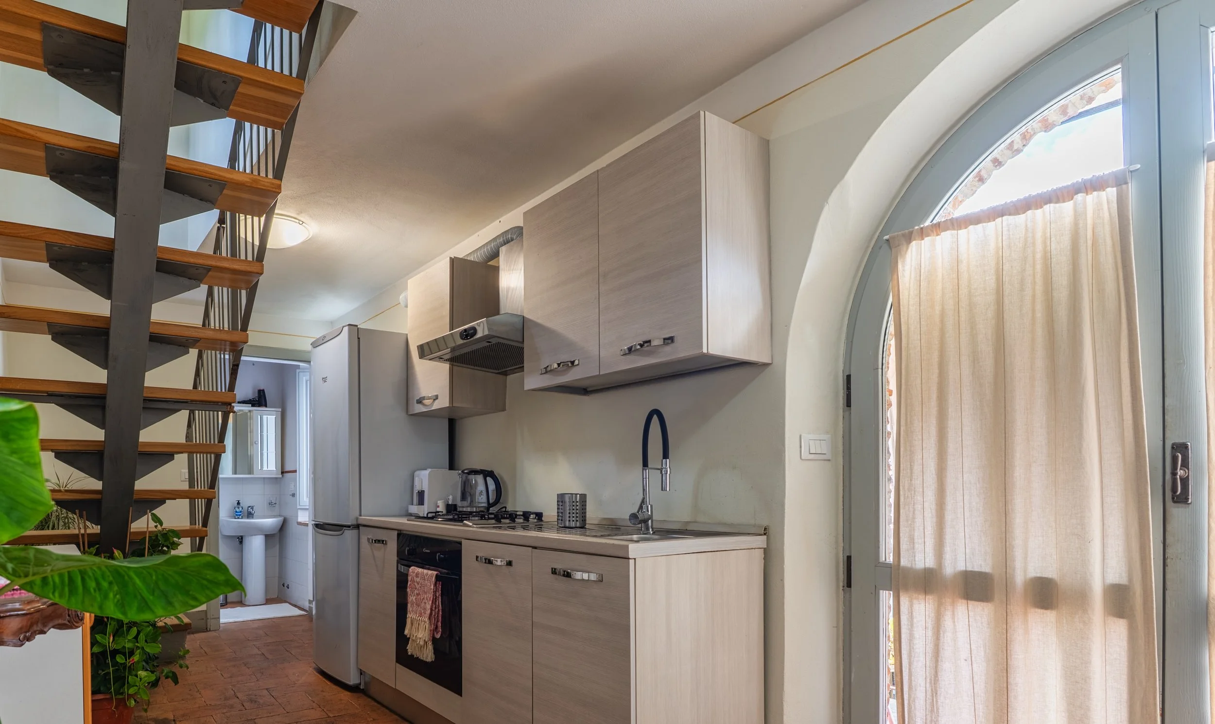 Interno di una cucina con mobili chiari, frigorifero, fornelli, forno, lavabo con rubinetto, e accessori da cucina, vicino a una porta con tenda beige e una scala di legno e metallo.
