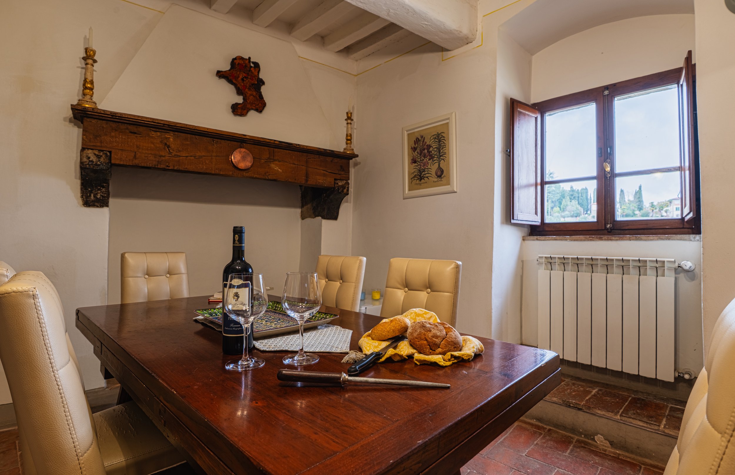 Sala da pranzo con tavolo in legno, quattro sedie imbottite beige, finestra aperta con vista su un giardino, pareti bianche, camino decorato con oggetti in terracotta e quadri alle pareti, bottiglia di vino, due bicchieri e pane su tovagliolo.