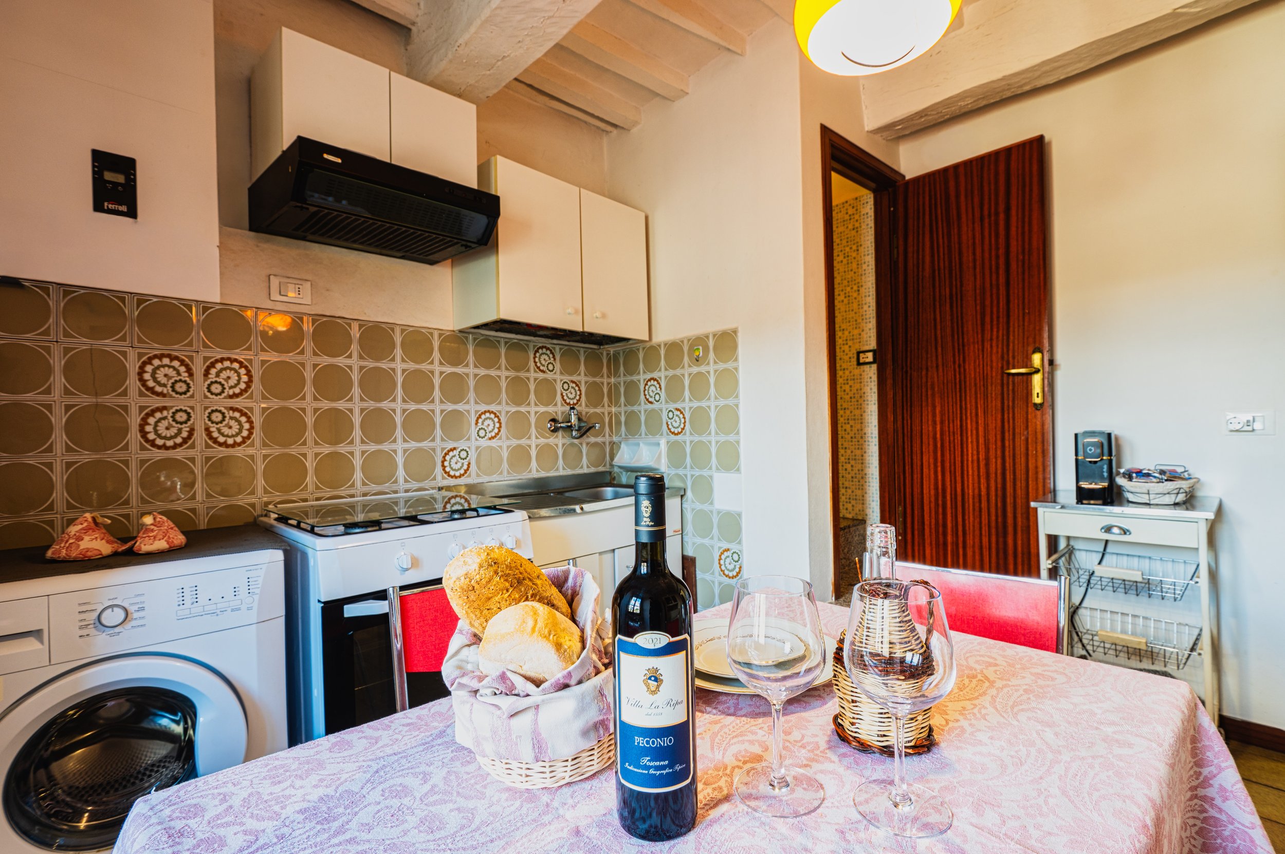 Cucina italiana con tavolo apparecchiato con pane, bottiglia di vino, bicchieri e decorazioni vintage, fornelli, cucina e dettagli d'epoca.