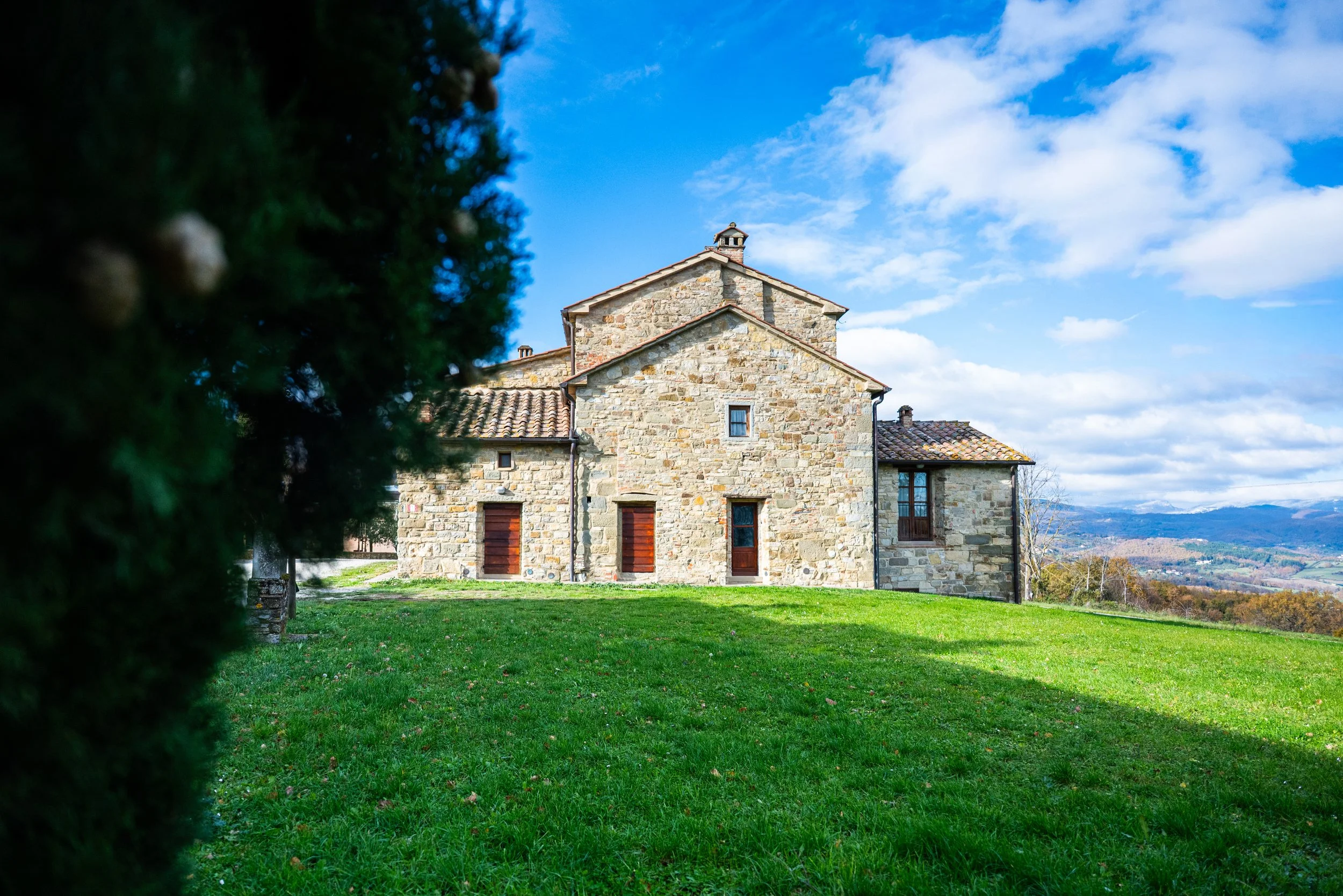 Casa in pietra con tetto di tegole in un paesaggio collinare, con cielo blu e nuvole bianche, circondata da un prato verde.
