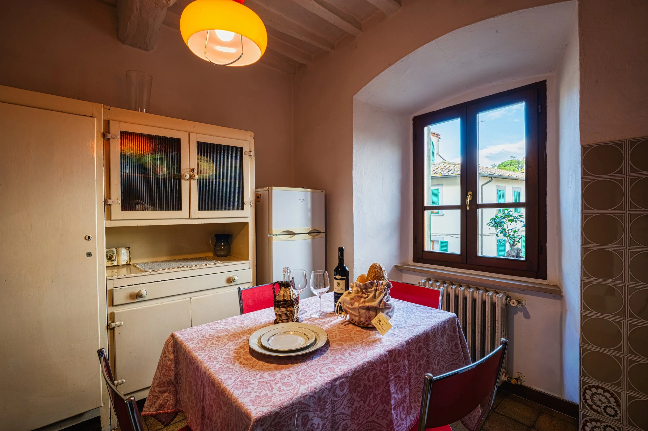 Una cucina rustica con tavolo apparecchiato con piatti, bicchieri e bottiglia di vino. Una finestra mostra case con persiane verdi, sotto un cielo parzialmente nuvoloso.