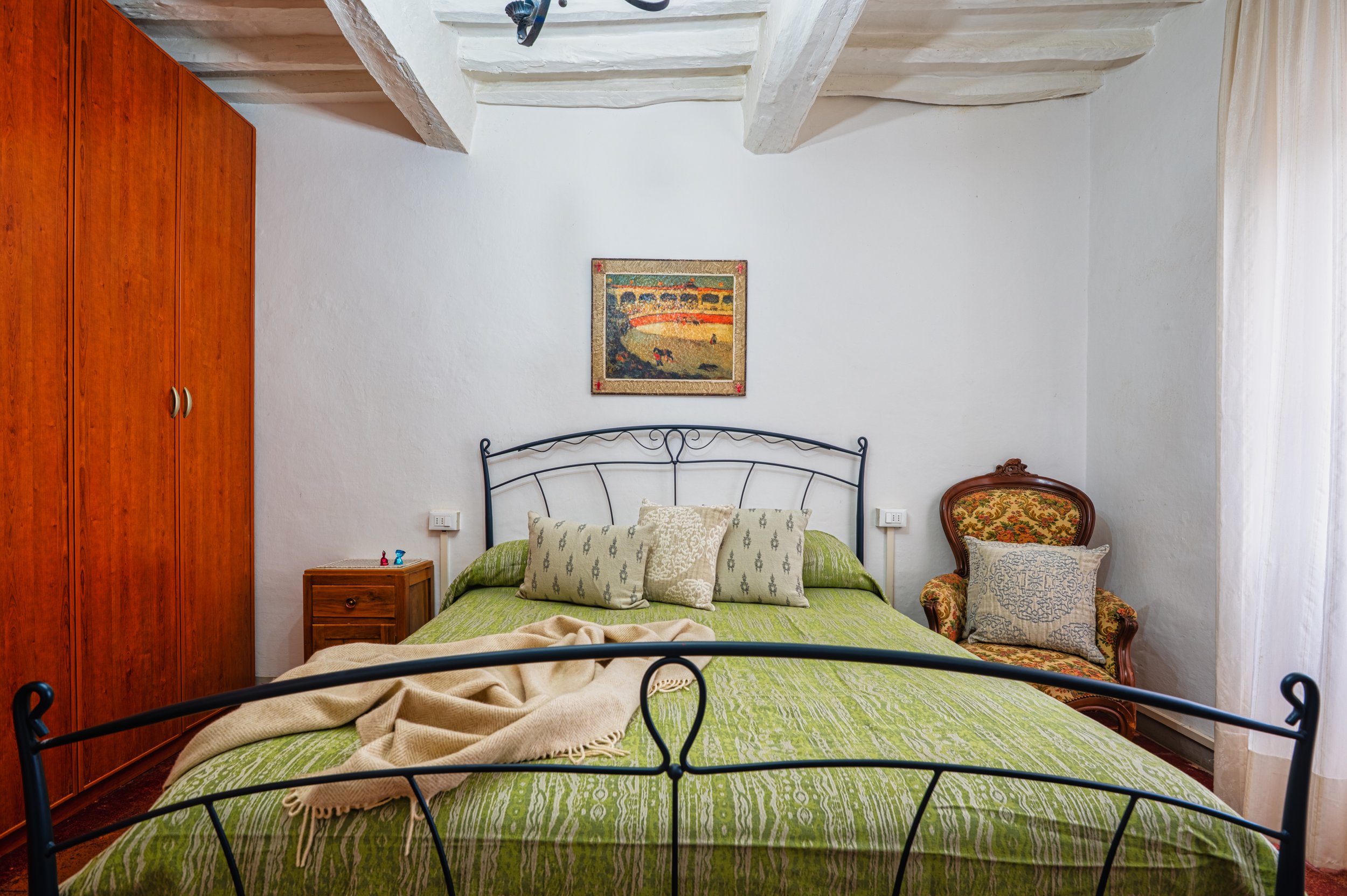 Camera da letto con letto matrimoniale, coperte verdi, quattro cuscini decorativi, armadio in legno a sinistra, piccolo comodino con alcuni oggetti, sedia imbottita con rivestimento floreale, quadro appeso al muro sopra il letto, pareti bianche, soff