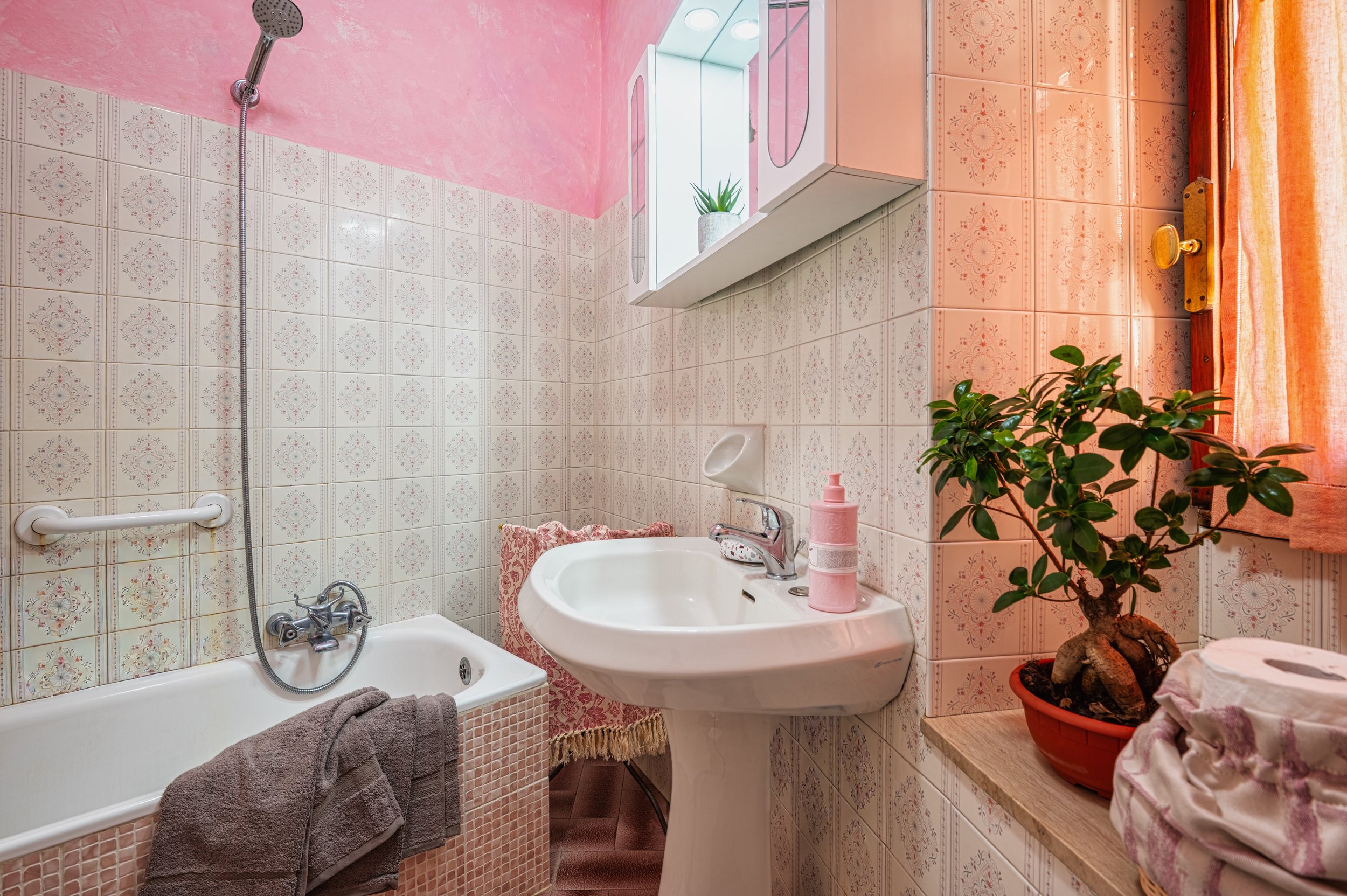 Bagno con lavabo, vasca da bagno, specchio con armadietto, piante in vaso, asciugamani, e tende alle finestre, decorato con piastrelle e pareti rosa.