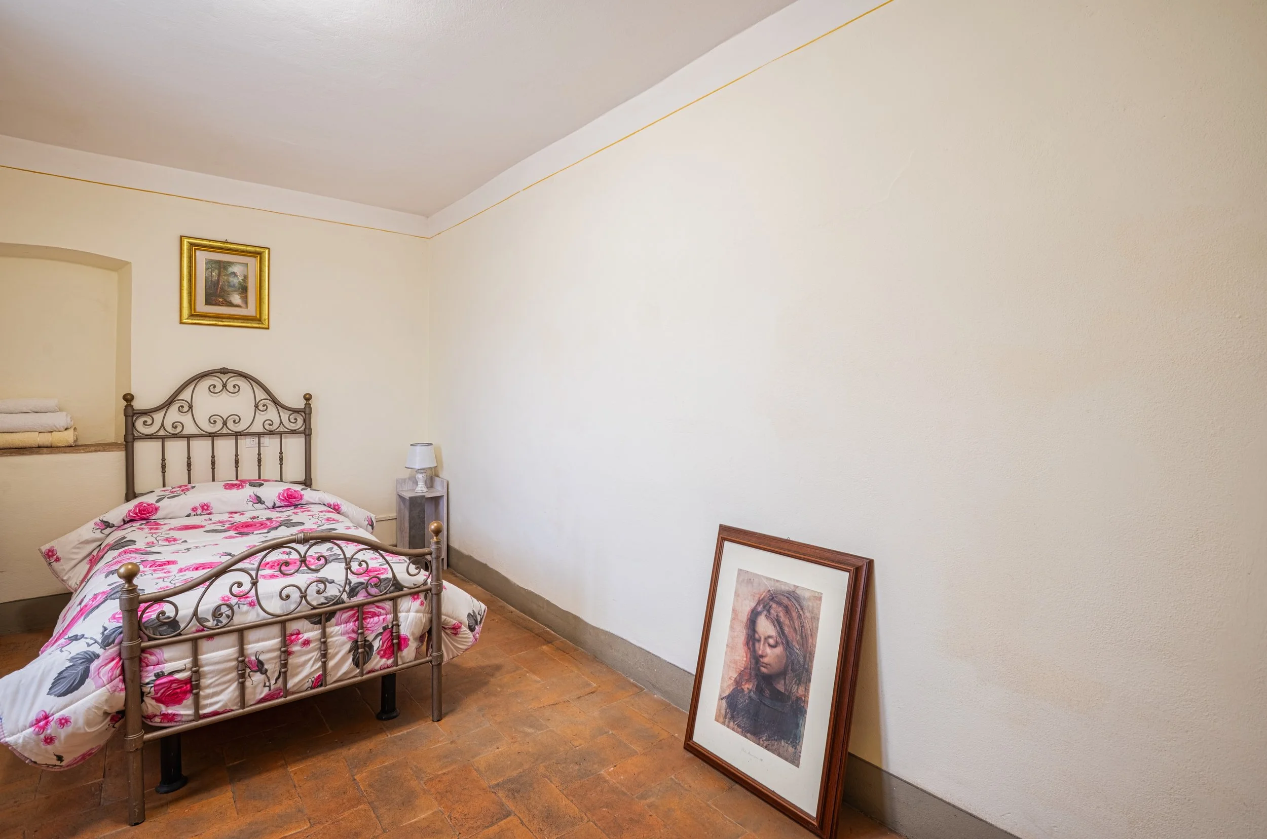 Camera con letto singolo in ferro battuto con copriletto decorato con rose, quadro con paesaggio sulla parete, poster di una donna con capelli lunghi appoggiato a terra, piccola lampada sul comodino.