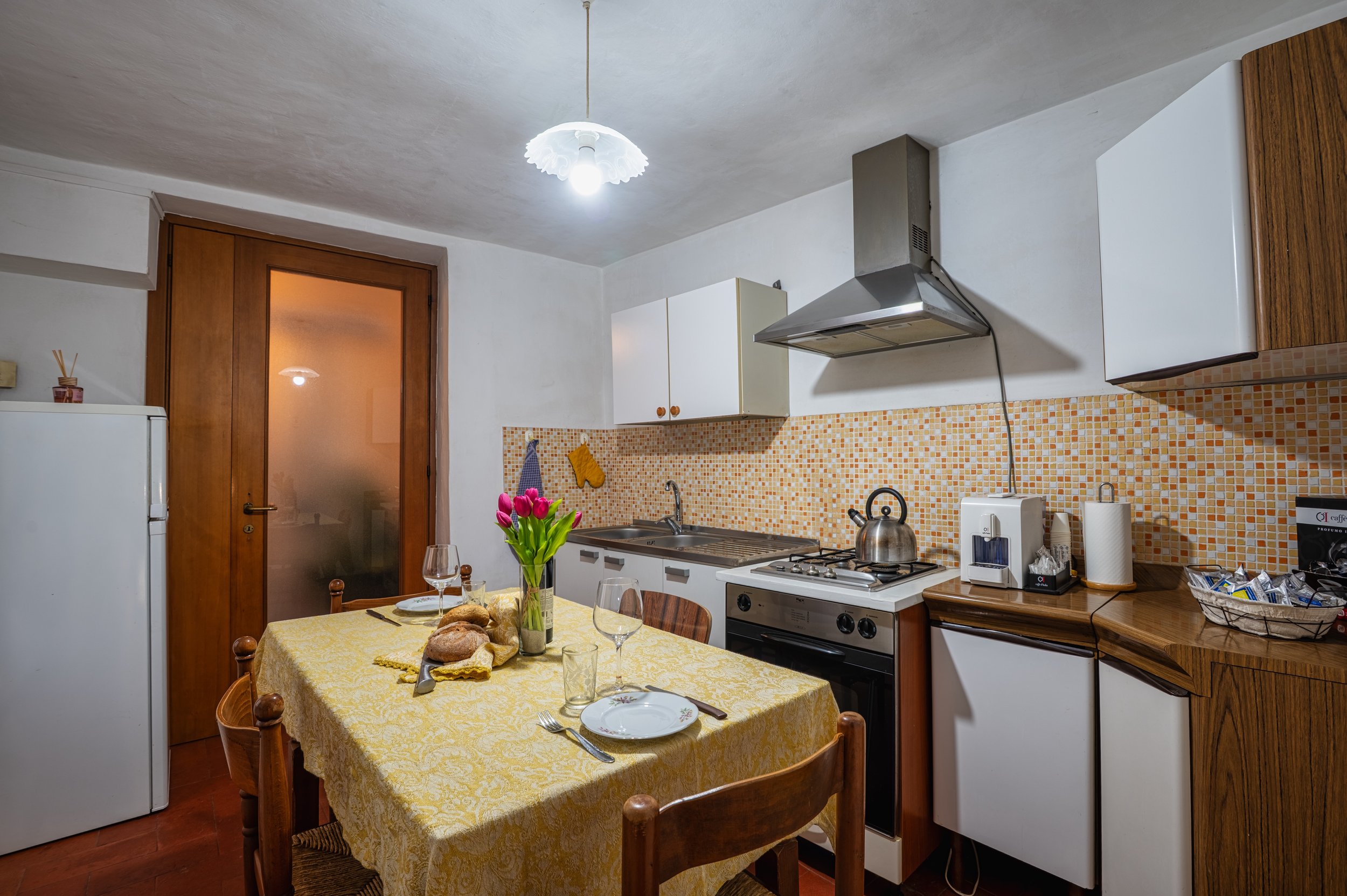 Cucina con tavolo ricoperto da tovaglia gialla, fiori rosa, pane, bicchieri e posate. Mobili in legno, cucina a gas, microonde, frigorifero e contemporanei accessori.