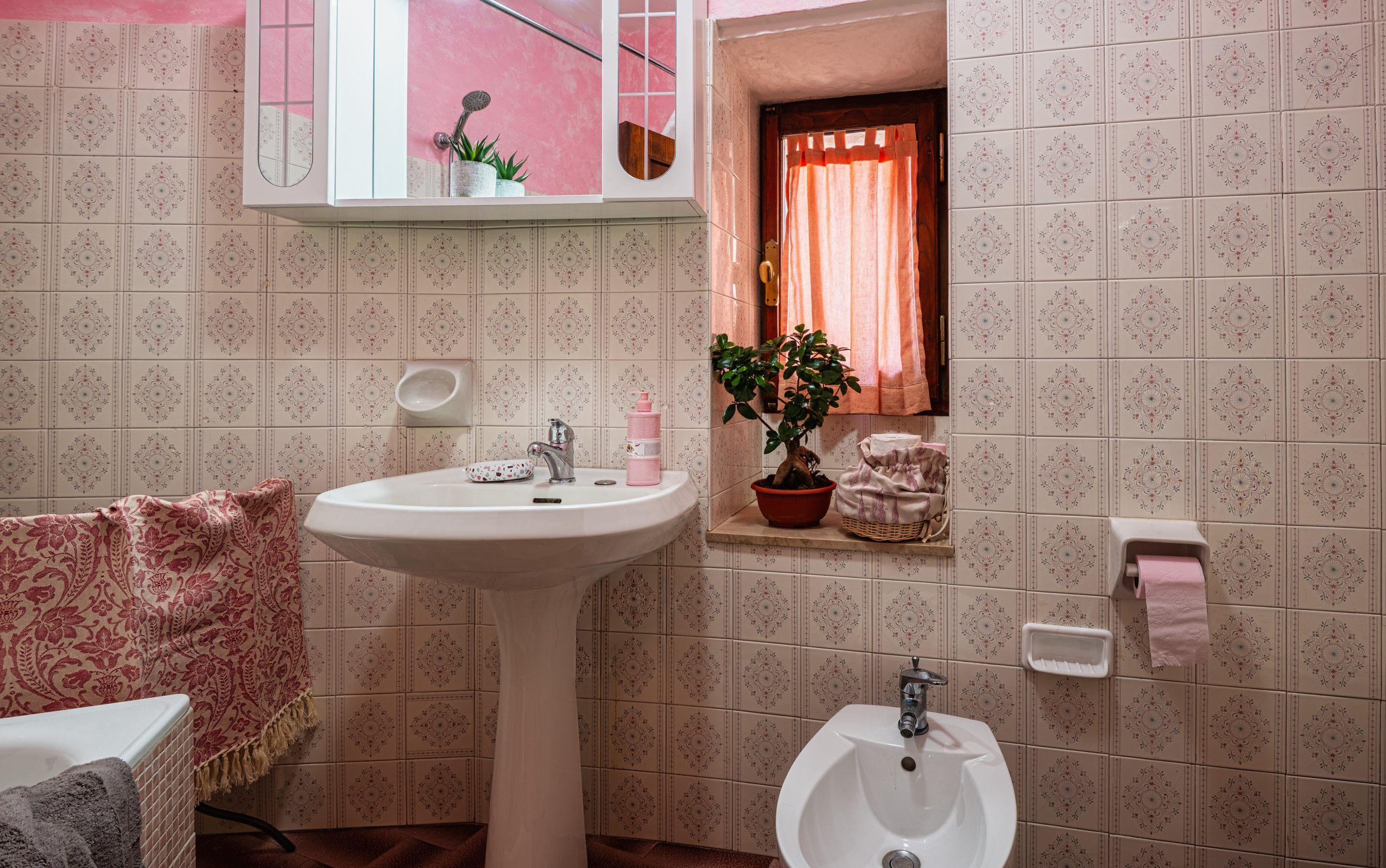 Bagno con pareti rivestite con piastrelle di motivo floreale, lavabo con doccetta, finestra con tende rosa, vaso di fiori, bidet, porta rotolo di carta igienica rosa, Specchio con mensola con piante verdi, rattan con tessuto e pianta sul davanzale