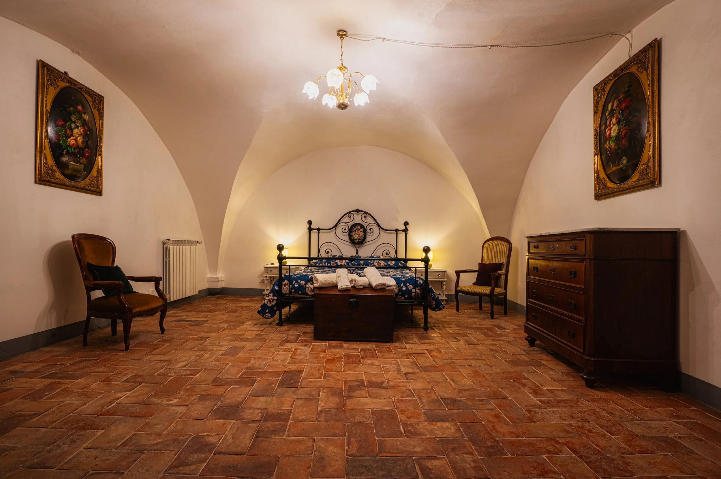 Camera da letto con soffitto a volta, letto in ferro battuto con biancheria floreale, due sedie di fronte al letto, armadio in legno scuro, quadri di fiori alle pareti, pavimento in terracotta, lampadario vintage.