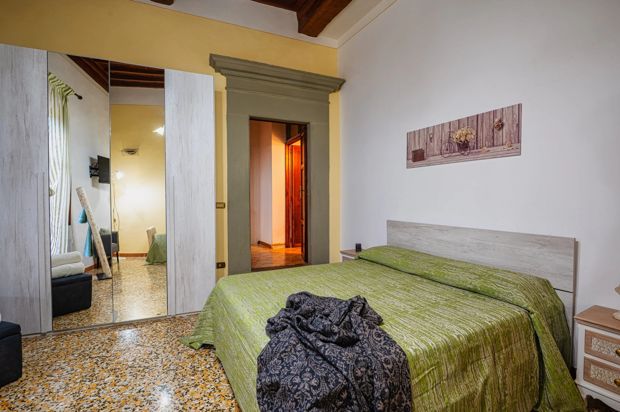 Camera da letto con letto matrimoniale, copriletto verde, armadio con specchi, pareti bianche e decorazioni rustic. Porta d'ingresso aperta e lampada accesa in soggiorno.