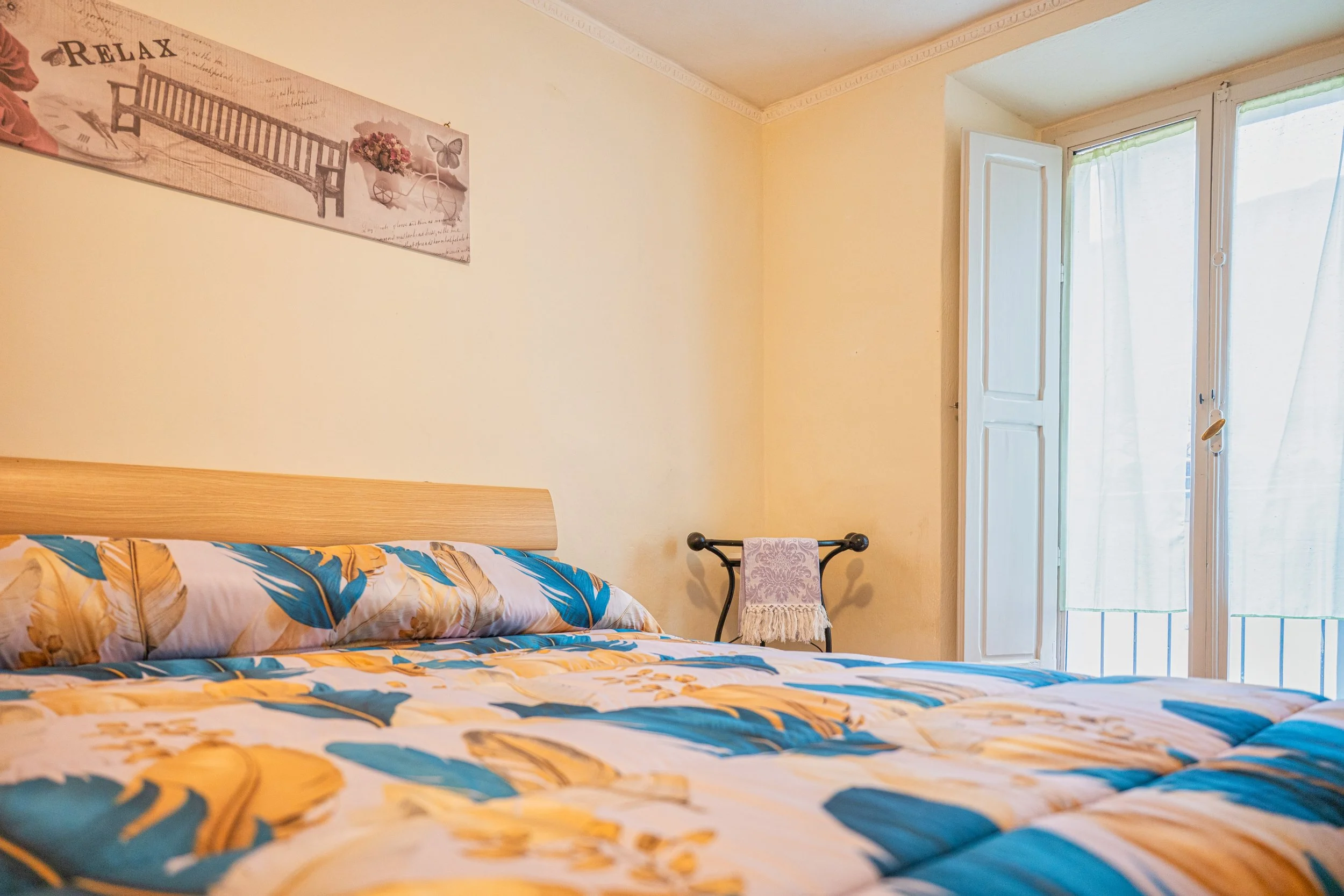 Camera da letto con lenzuola a motivi floreali gialli e blu, finestra con tende bianche e porta aperta che dà su un balcone, quadro decorativo sulla parete con la scritta "Relax".