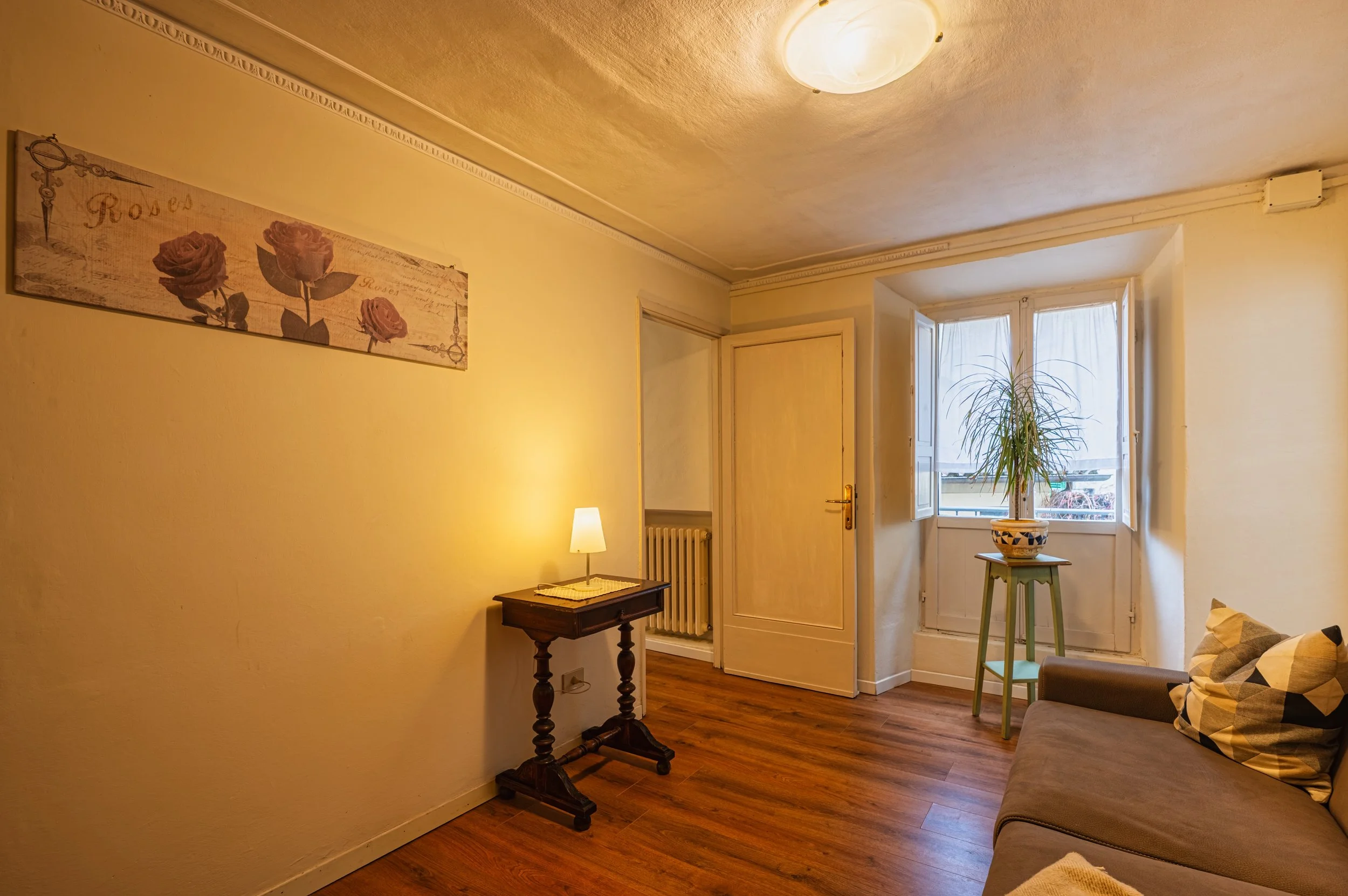 Interno di un soggiorno con pareti gialle, un divano beige con cuscini, un tavolino in legno con una lampada accesa, una finestra aperta con tende bianche e una pianta in un vaso sopra un supporto verde.