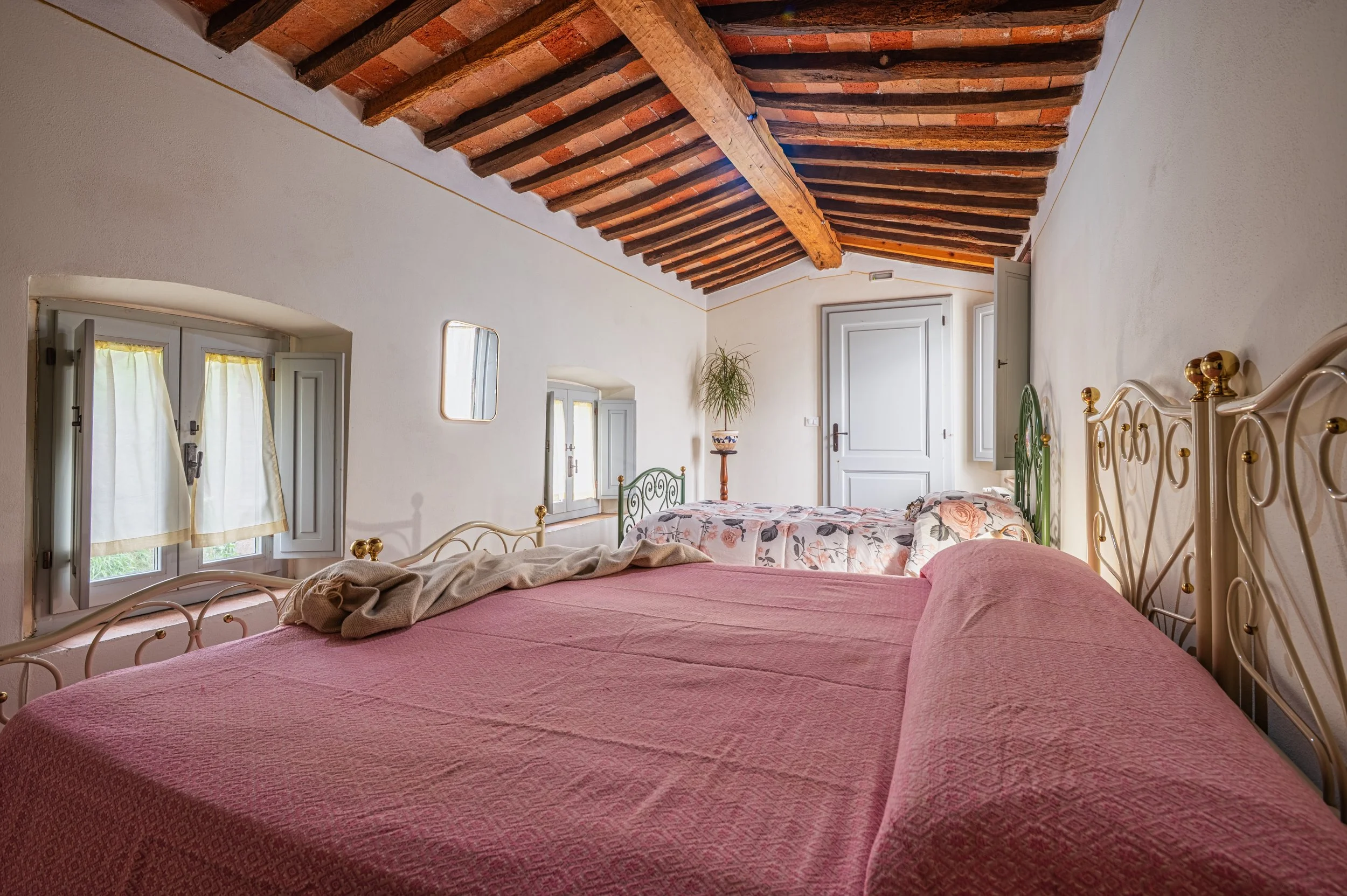 Camera da letto con soffitto in legno e pareti bianche, due letti con testiere in metallo decorato, uno con copriletto rosa e l'altro con copriletto con motivo floreale, finestre con tende leggere, una pianta in vaso su un tavolino in fondo alla stan