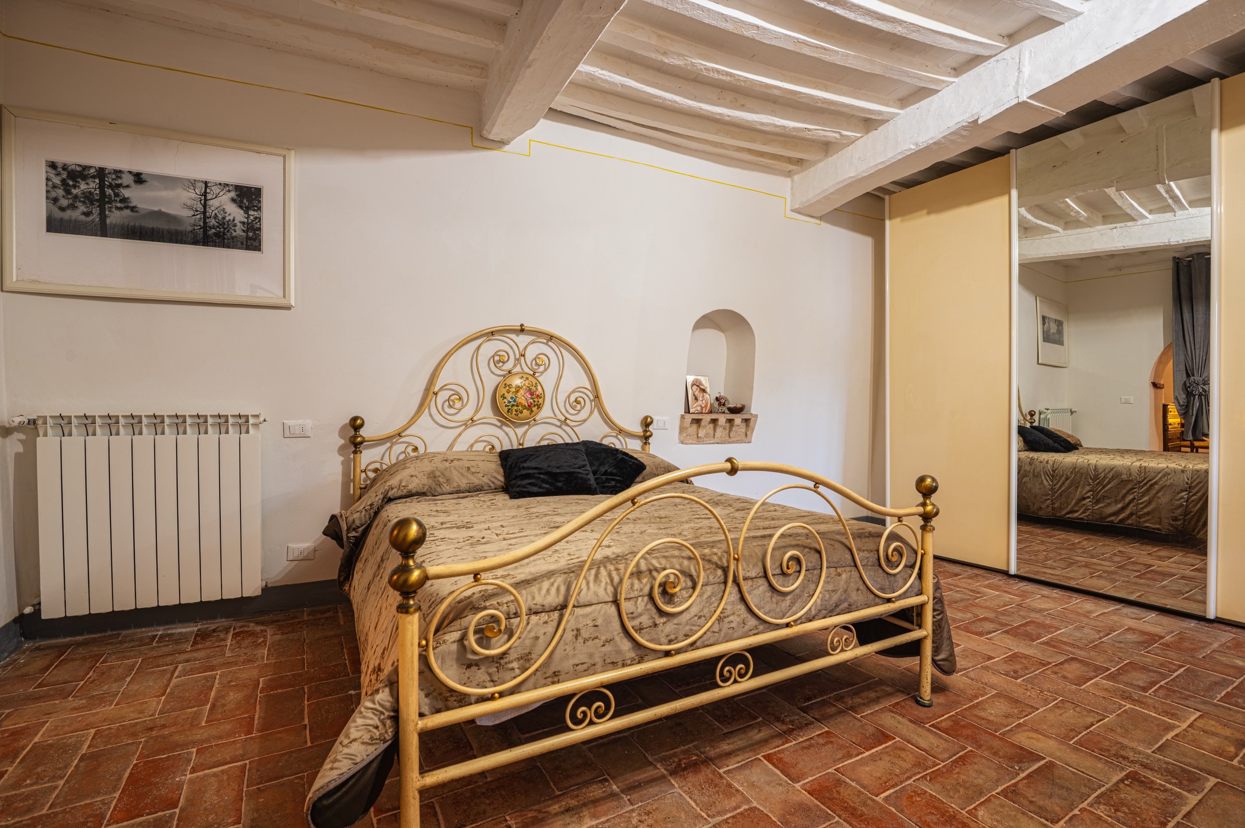 Camera da letto con letto in ferro battuto dorato, pareti bianche, pavimento in piastrelle di terracotta, specchio a specchio grande con mobile in fondo, dipinto con alberi e nebbia, nicchia con decorazioni, porta scorrevole con specchio che riflette