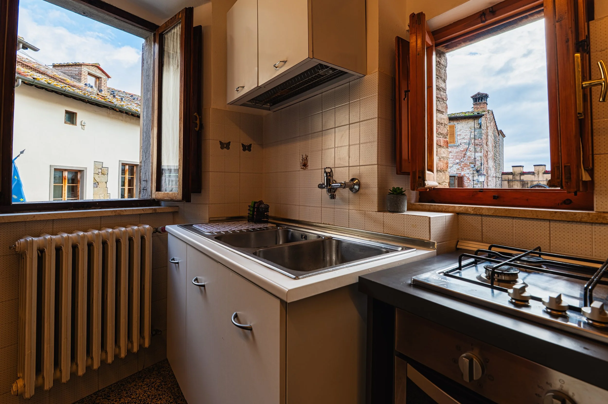 Cucina con due finestre aperte che mostrano edifici circostanti, con lavabo e piano cottura a gas