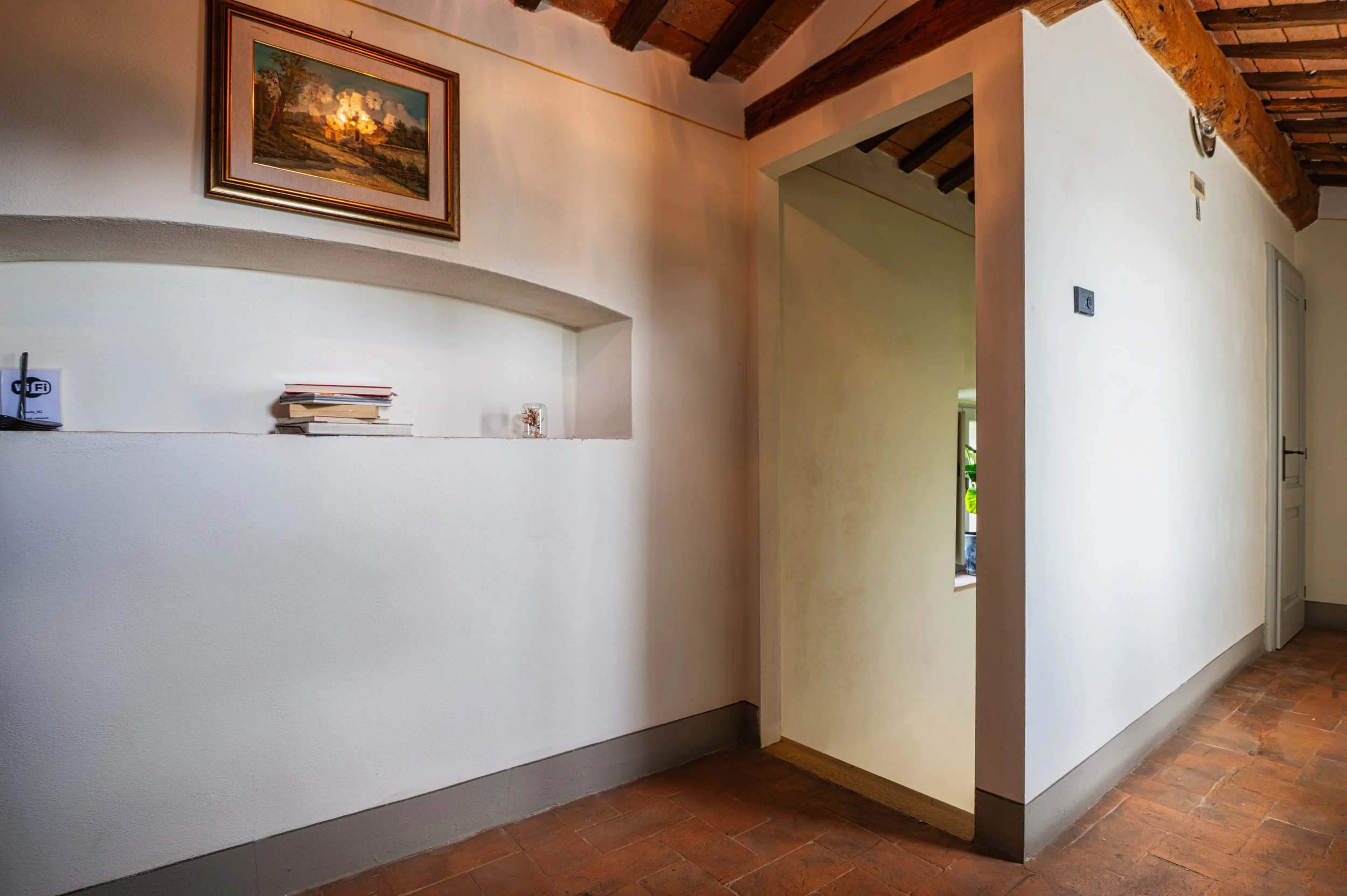 Interno di una casa con pareti bianche, soffitto con travi di legno, pavimento in piastrelle di terracotta, una nicchia con alcuni libri e decorazioni, quadro appeso alla parete, porte e finestre che lasciano entrare luce naturale.