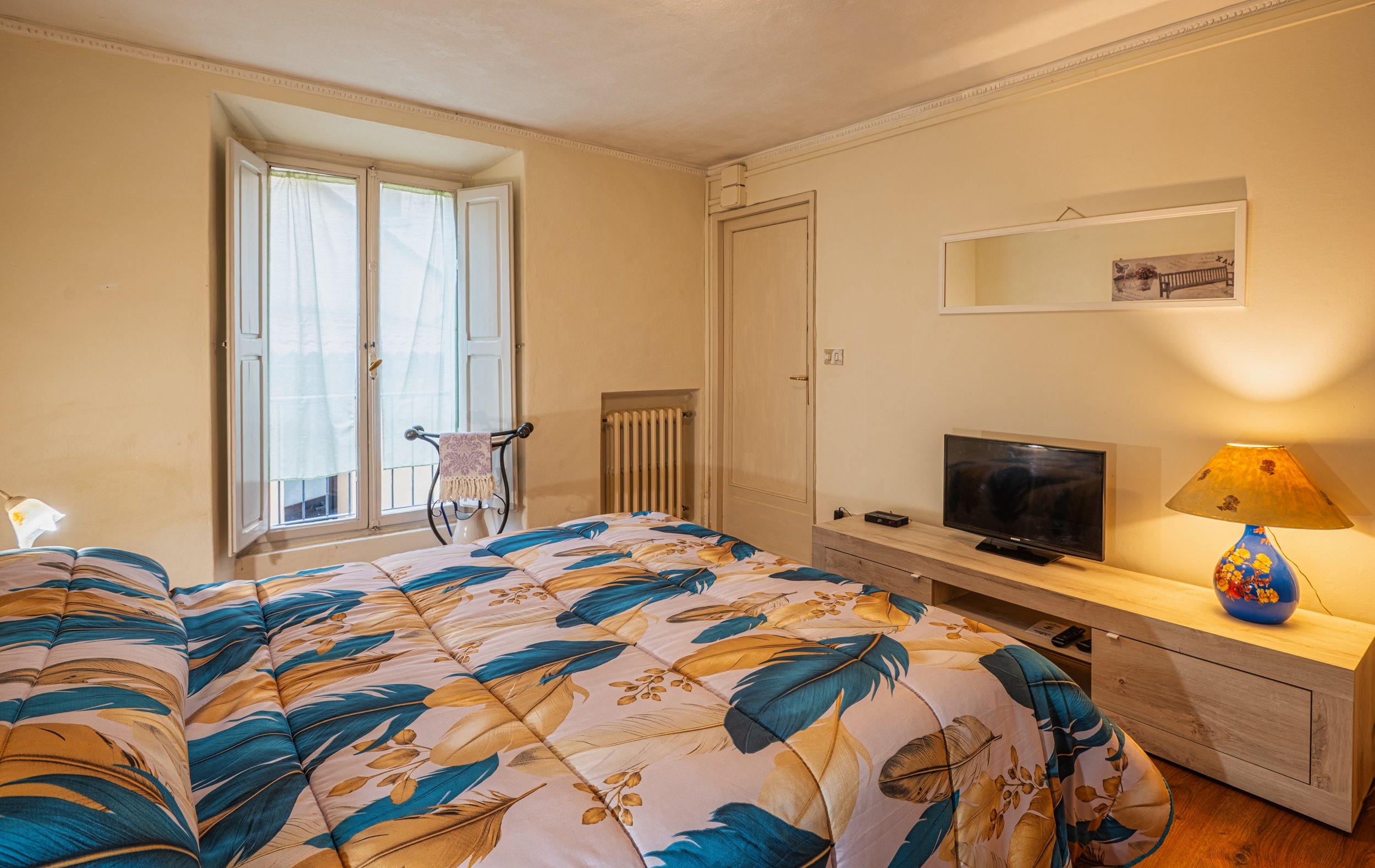 Una camera da letto con un letto matrimoniale con copripiumino dai motivi floreali blu e beige. È presente un televisore su una mensola in legno chiaro, una lampada con paralume colorato e una finestra con persiane socchiuse e tende leggere.
