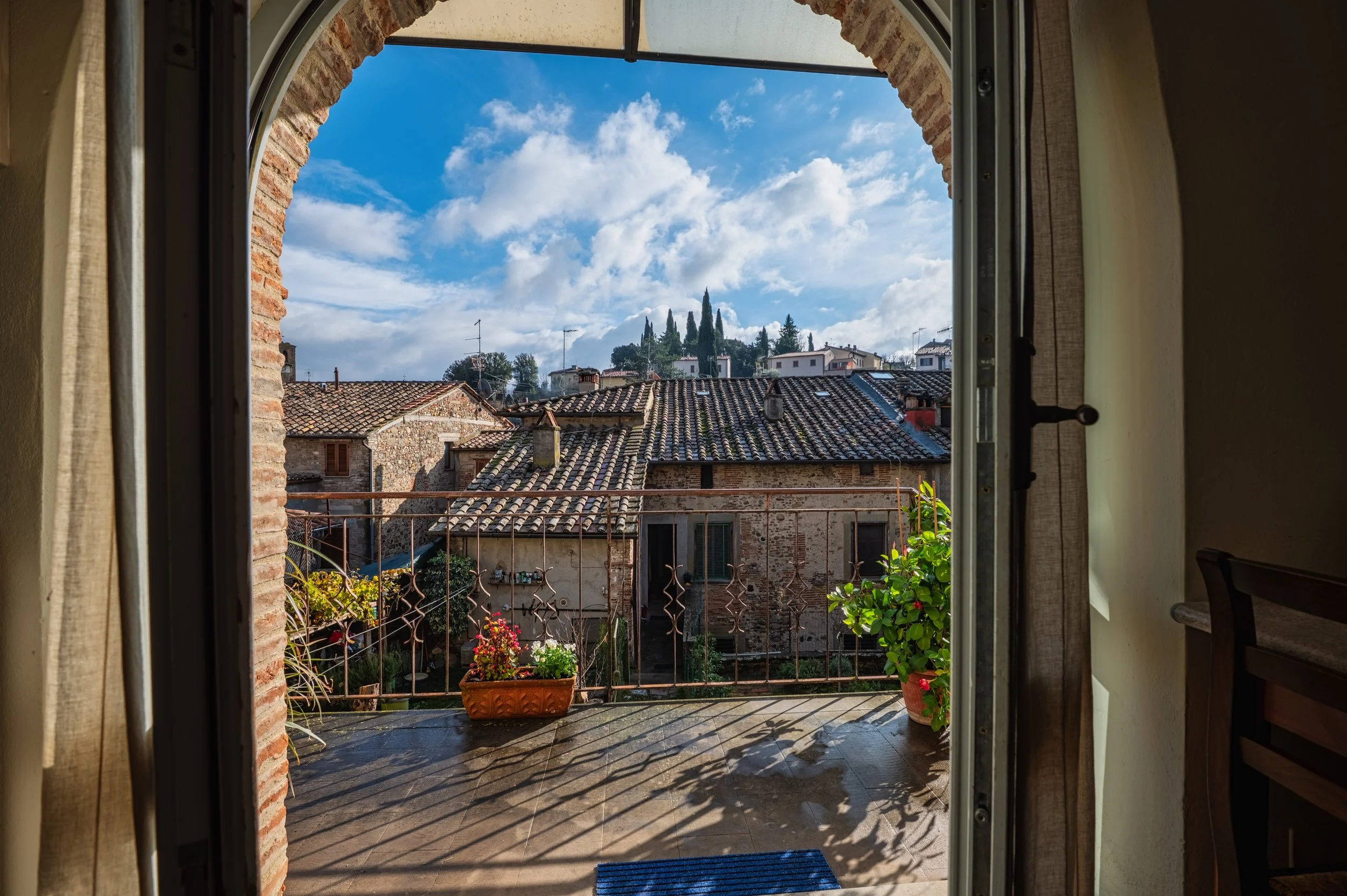 Vista da un balcone con piante e fiori, su tetti di tegole in un borgo italiano, con cielo nuvoloso e alcuni alberi sullo sfondo.