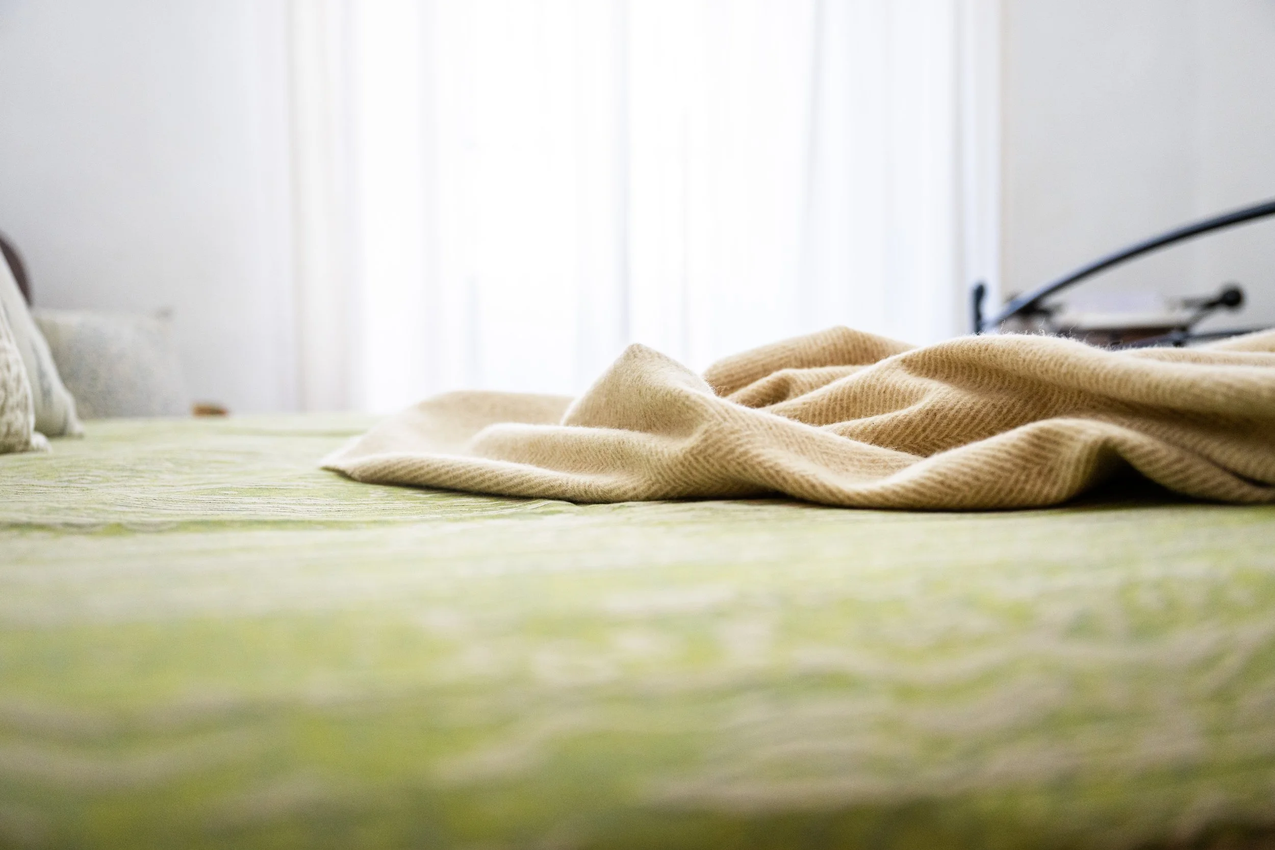 Letto con lenzuola di colore beige e coperta piegata, con cuscini ai lati, sotto una finestra con tende bianche leggere.