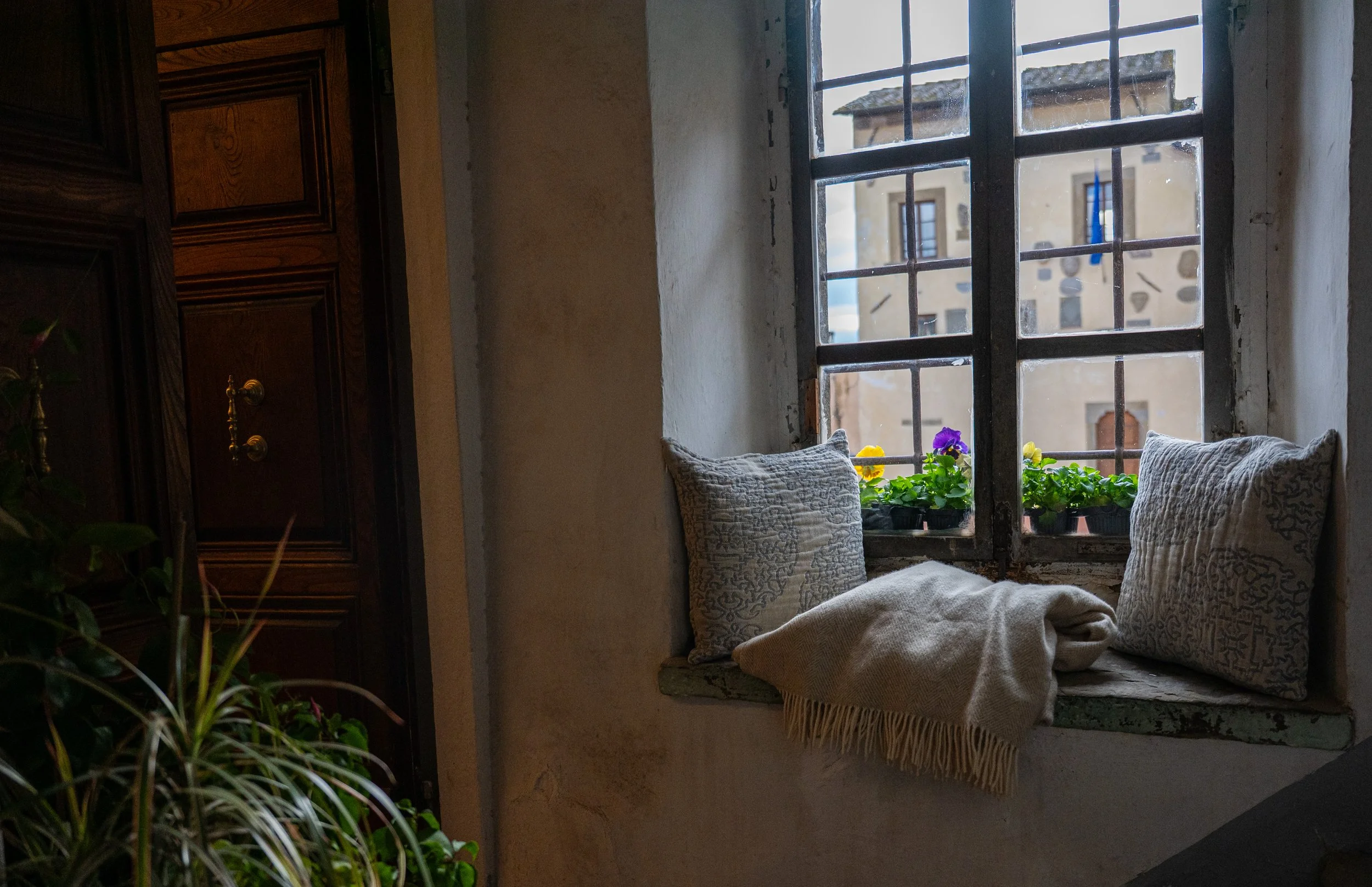 Interno di una stanza con una finestra con sbarre, sedile decorato con cuscini e pantofole, e piante con fiori alla finestra.