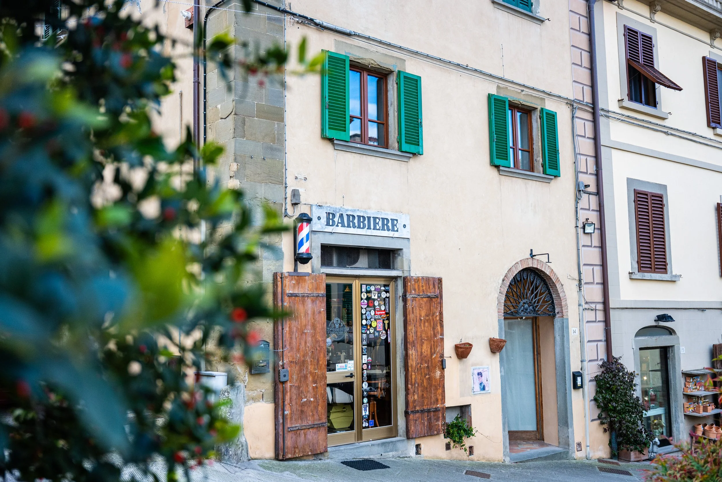 Vicolo con negozio di barbiere chiamato 'Barbiere', con finestra e porta antica, decorazioni floreali, piante e case con persiane verdi e marroni.
