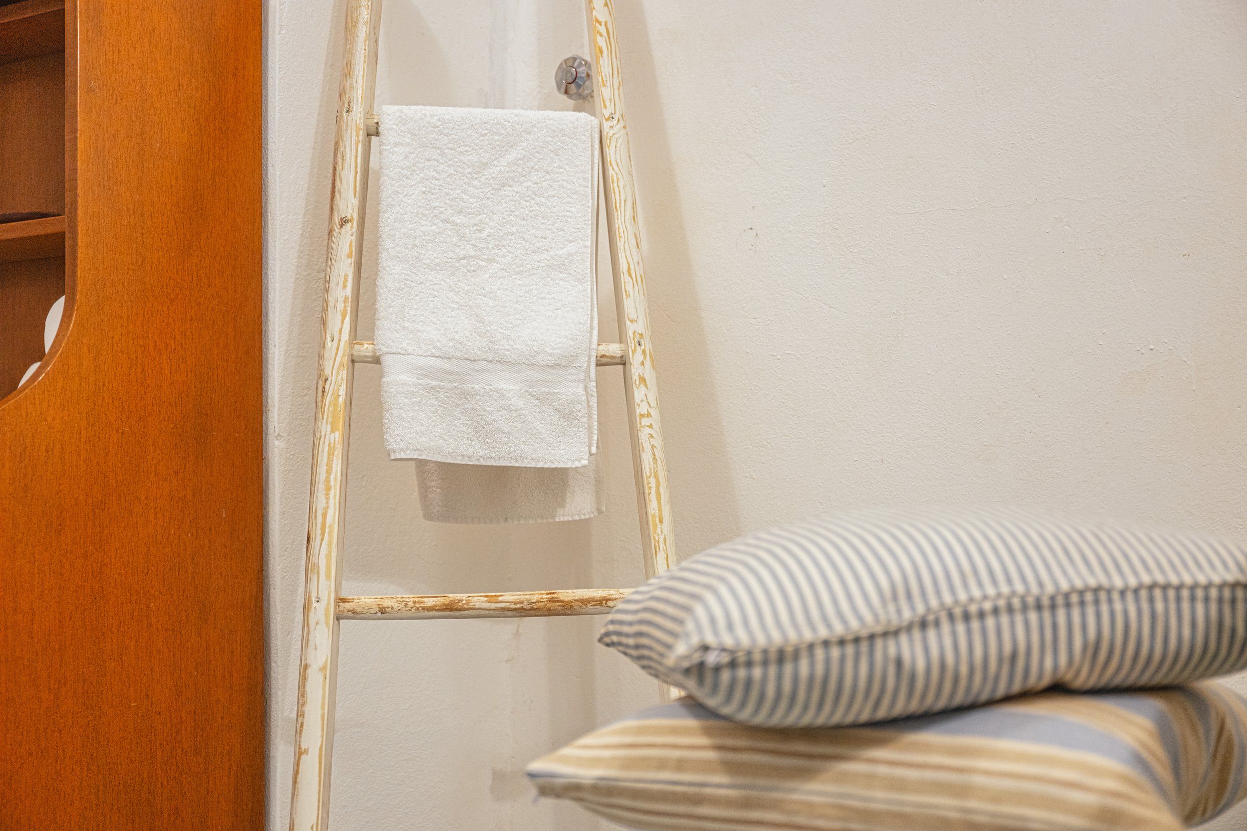 Una sedia con un cuscino a righe e un altro cuscino sotto, un asciugamano bianco su una scaletta di legno e un muro beige sullo sfondo.