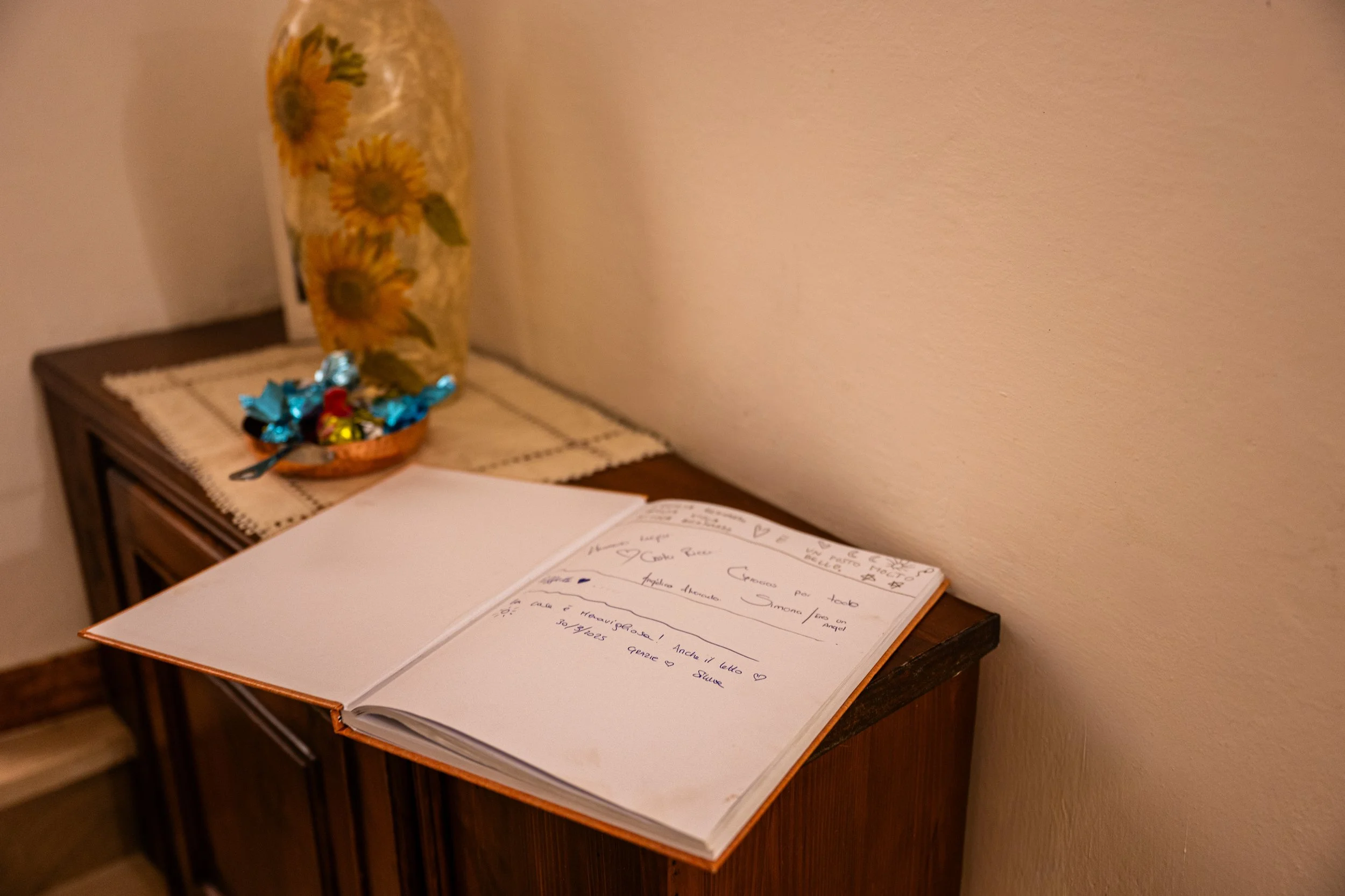 Libro aperto con scritte e disegni sulla pagina, posato su un mobile di legno. Sullo sfondo, un vaso con fiori di girasole e un piattino con cioccolatini avvolti in carta colorata, sopra una tovaglietta beige.