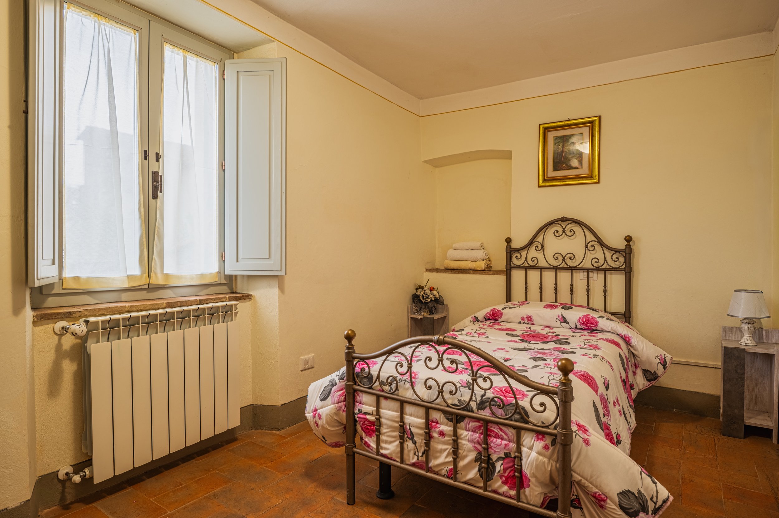 Camera da letto con finestra con tende bianche, letto con copertura floreale rosa e grigia, cuscini sulla testiera, quadri sulle pareti e arredamento semplice.